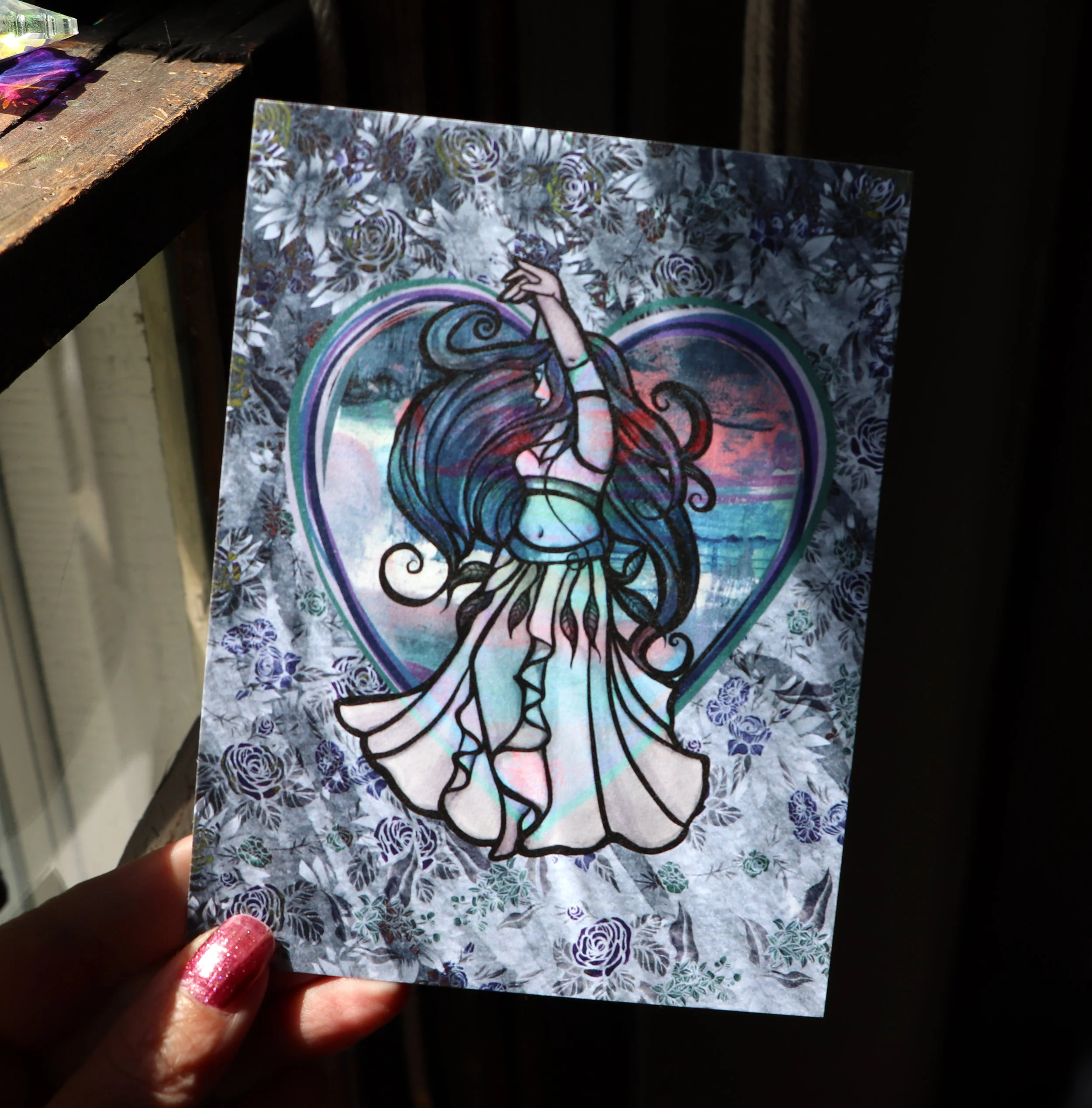 A person with pink nail polish holding a decorative card featuring an intricate line drawing of a woman with flowing hair, floral patterns, and rainbow colors.