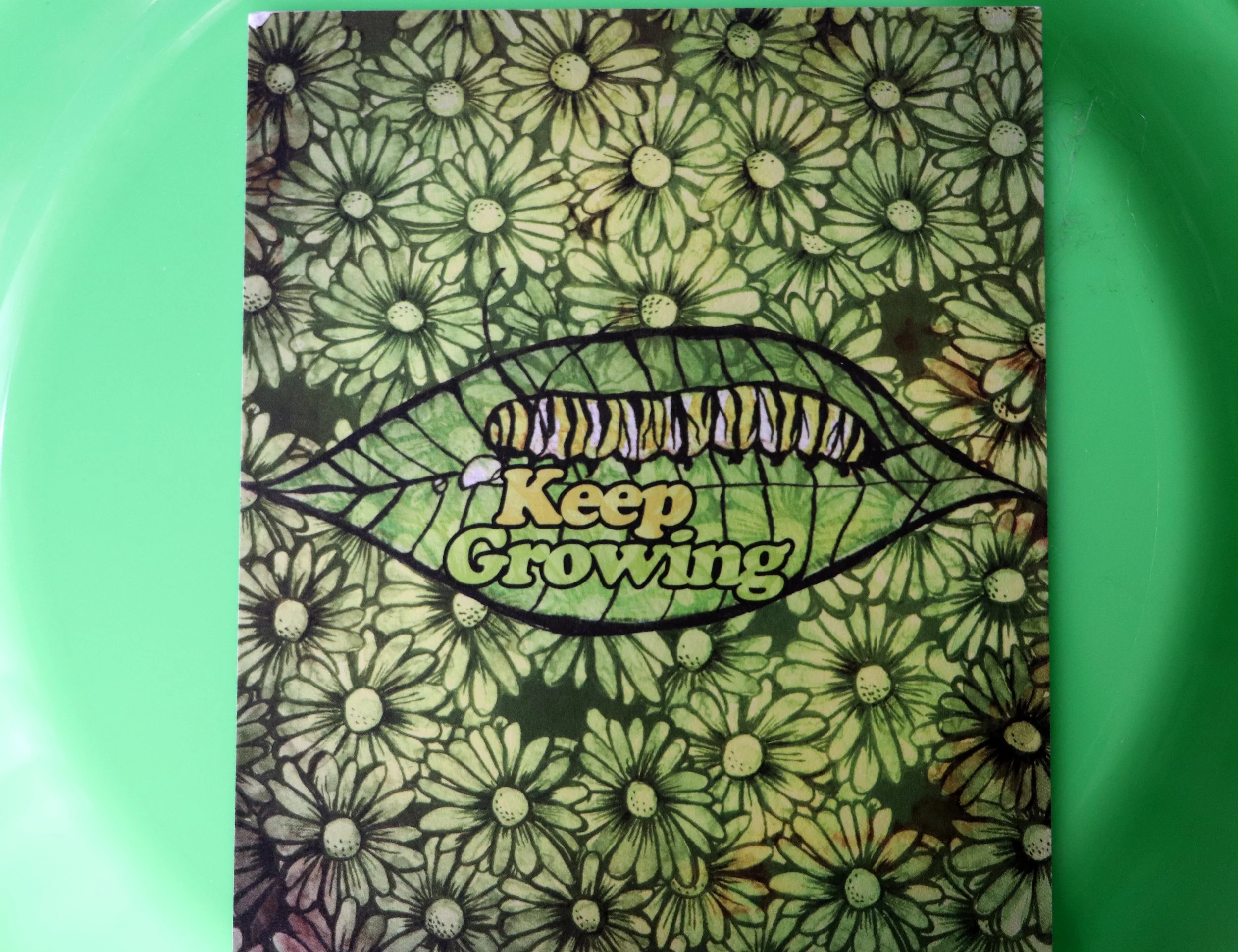 A book cover titled 'Keep Growing' with black outline drawings of daisies on a yellow-green background, partly covered by a bright green surface.