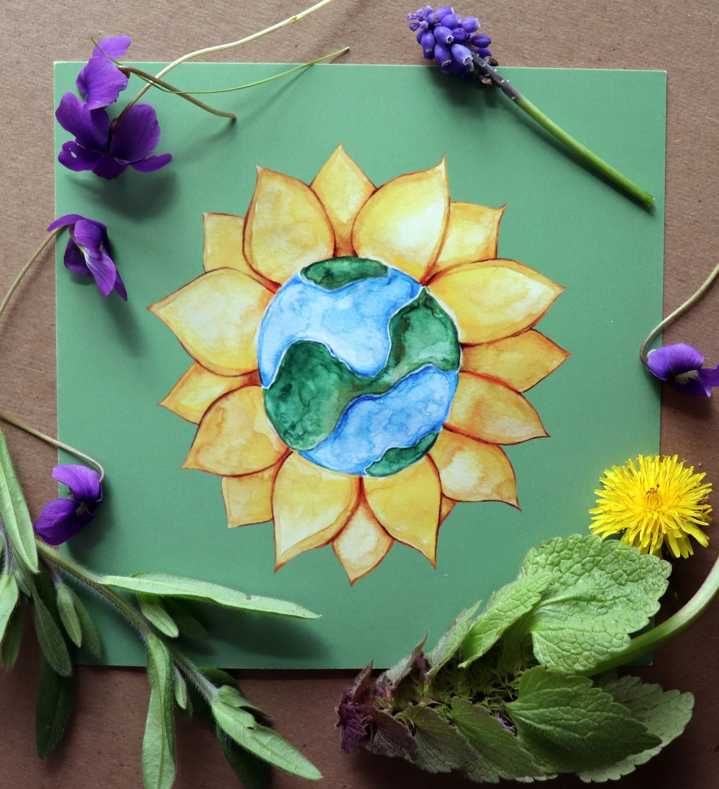 A drawing of a sunflower with a blue and green Earth globe at its center, surrounded by various real flowers and green leaves.