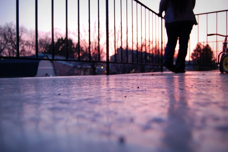 A person walking on a concrete surface near a red metal fence during sunset, with the person's silhouette visible and a bicycle nearby.