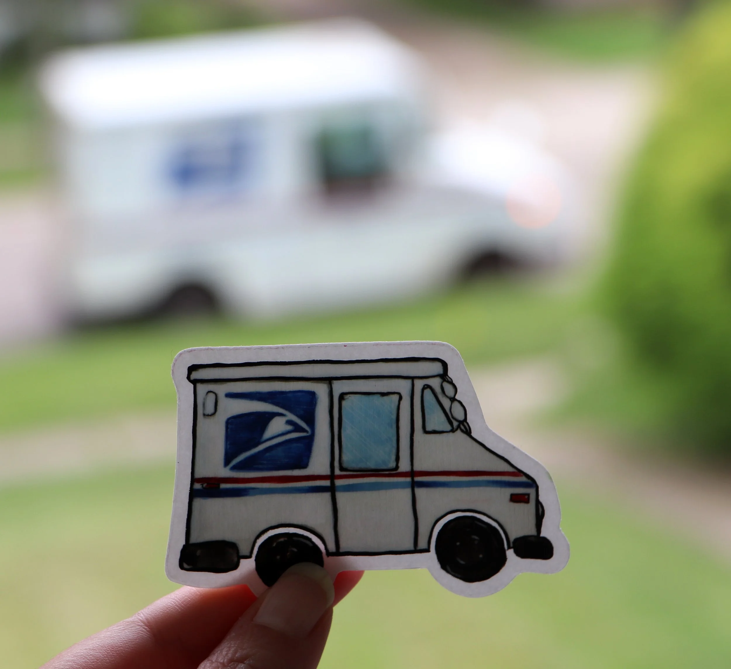 Hand holding a paper cutout of a USPS mail truck in front of a blurred USPS truck outside.