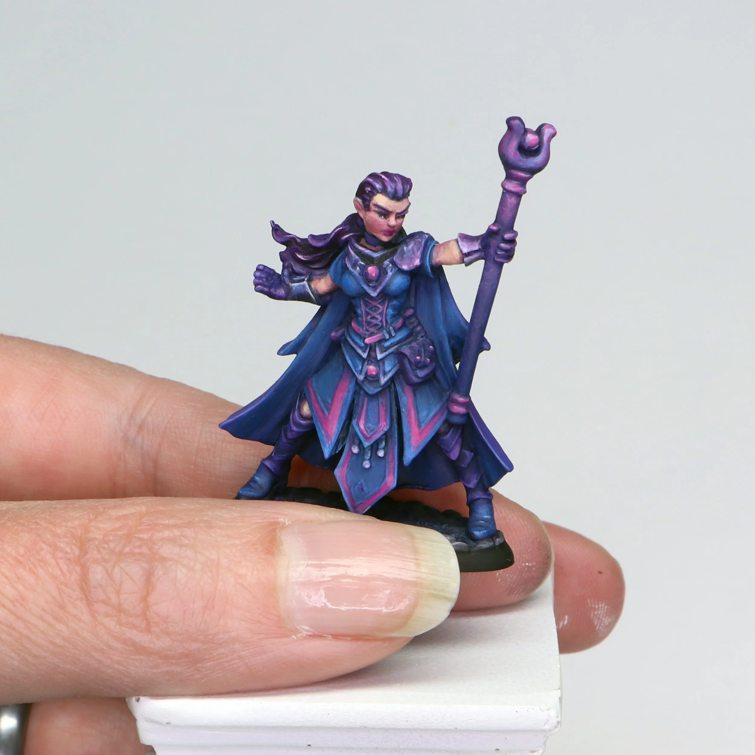 Close-up of a small, painted figurine of a female character dressed in purple and blue, holding a staff, standing on a finger against a light background.