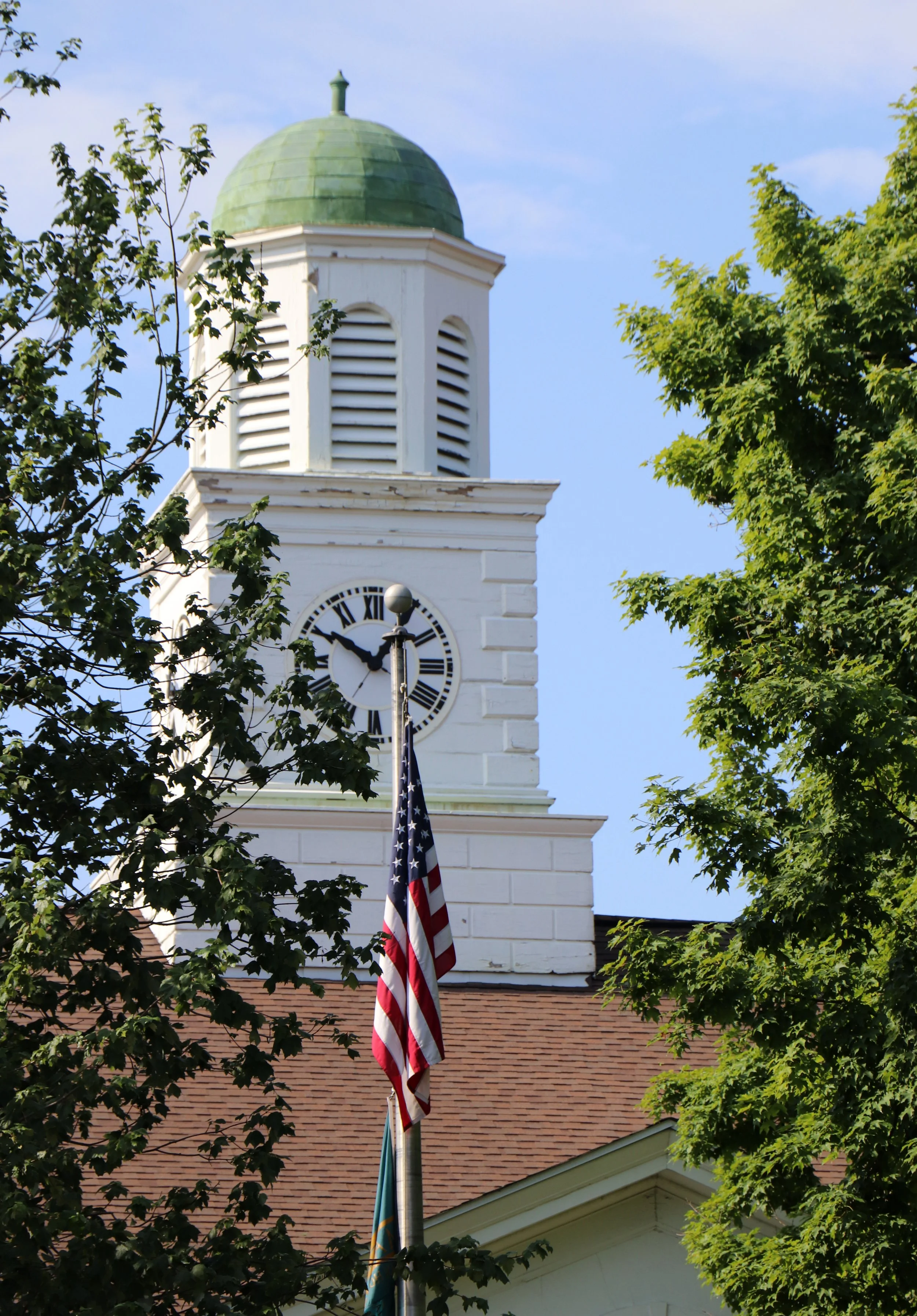 View of a white clock tower with a green dome, framed by leafy green trees, with an American flag hanging from a flagpole in front.