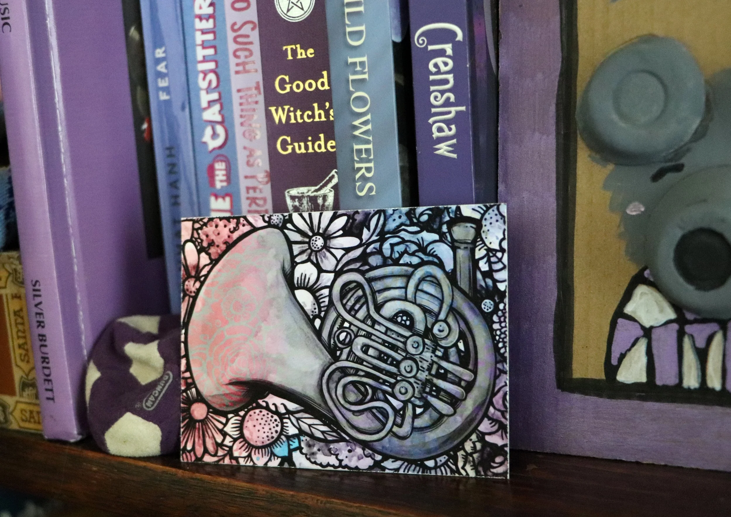 A shelf with purple books about creatures, witches, and fantasy, and a colorful greeting card featuring a black and white illustration of a French horn among flowers, with some purple and gray objects nearby.