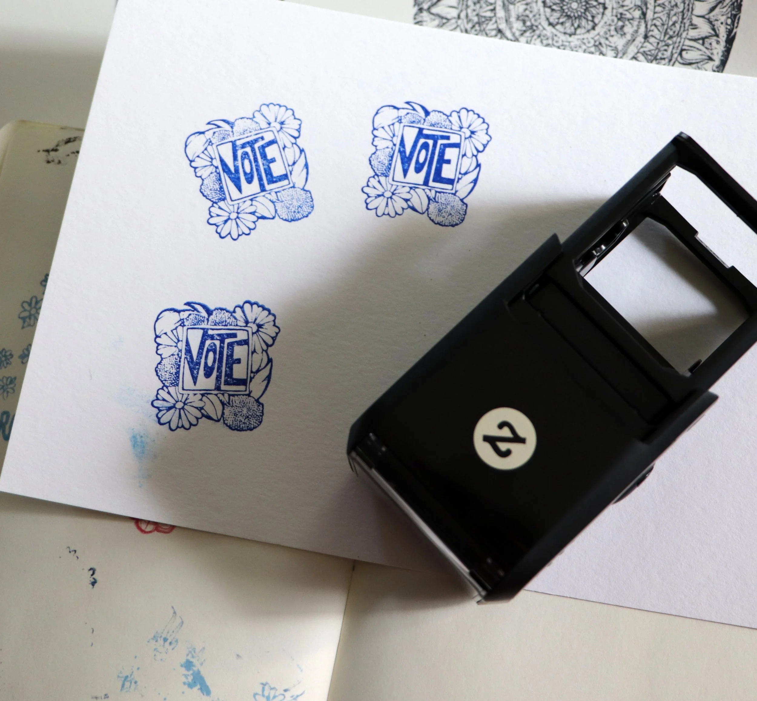 A sheet of paper with three blue 'VOTE' stamps featuring floral designs, and a black stamp maker positioned on the paper.
