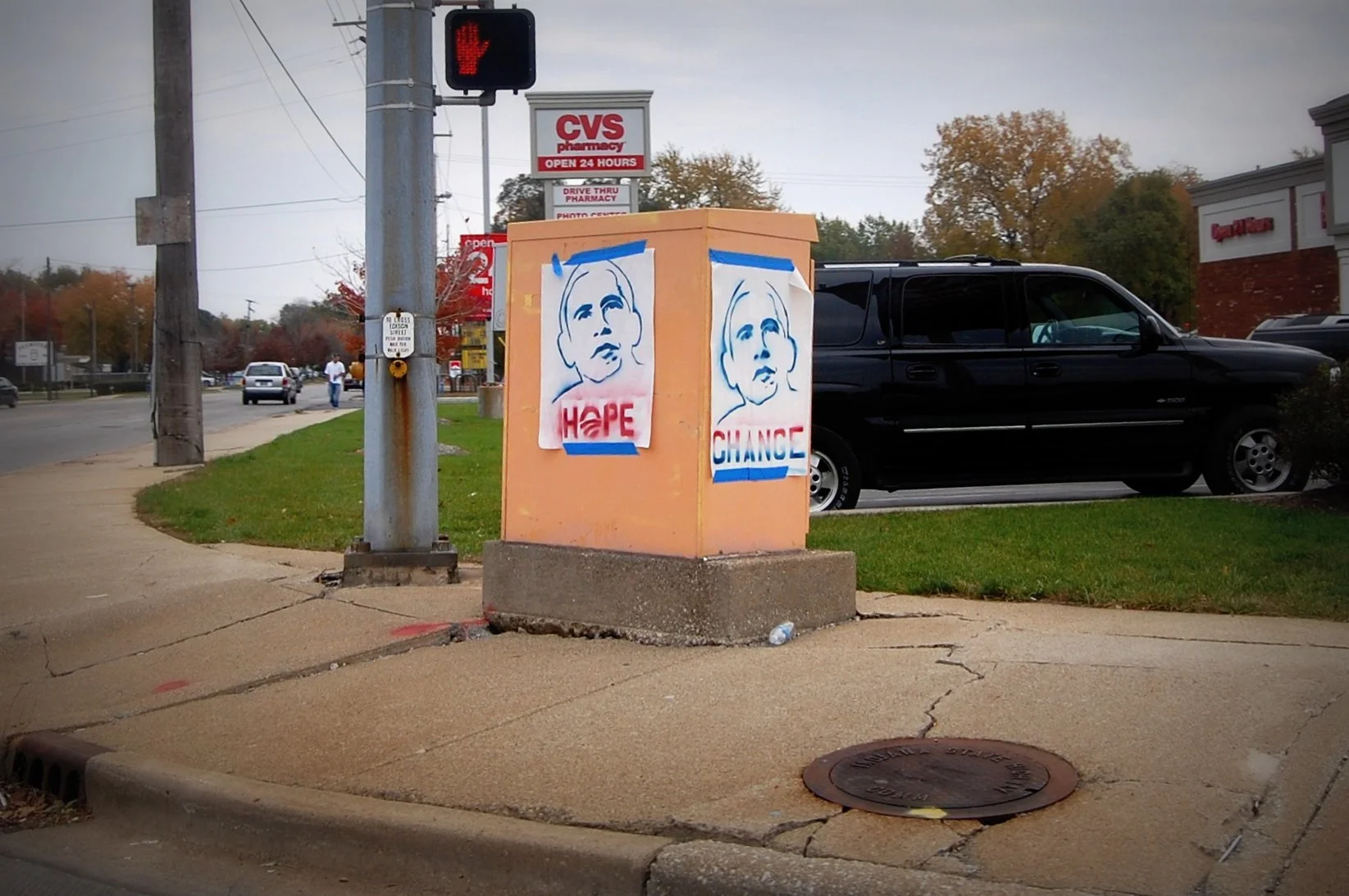 Street corner with political posters, traffic light showing red hand, CVS pharmacy sign in background, parked black SUV, and sidewalk with cracks.
