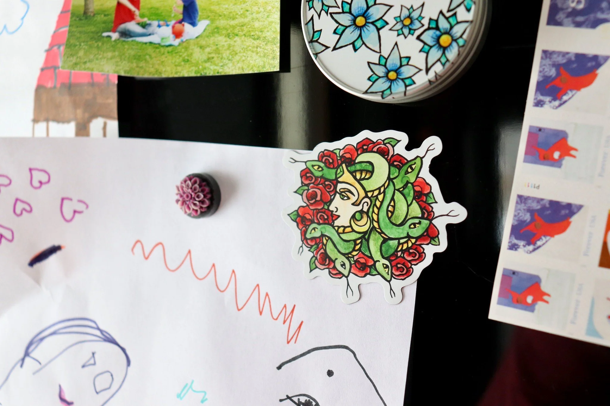 Various colorful drawings, stickers, and photographs are attached to a surface. Notable items include a sticker of Medusa with snakes for hair, a photograph of people picnicking, and decorative stickers with floral and animal motifs.
