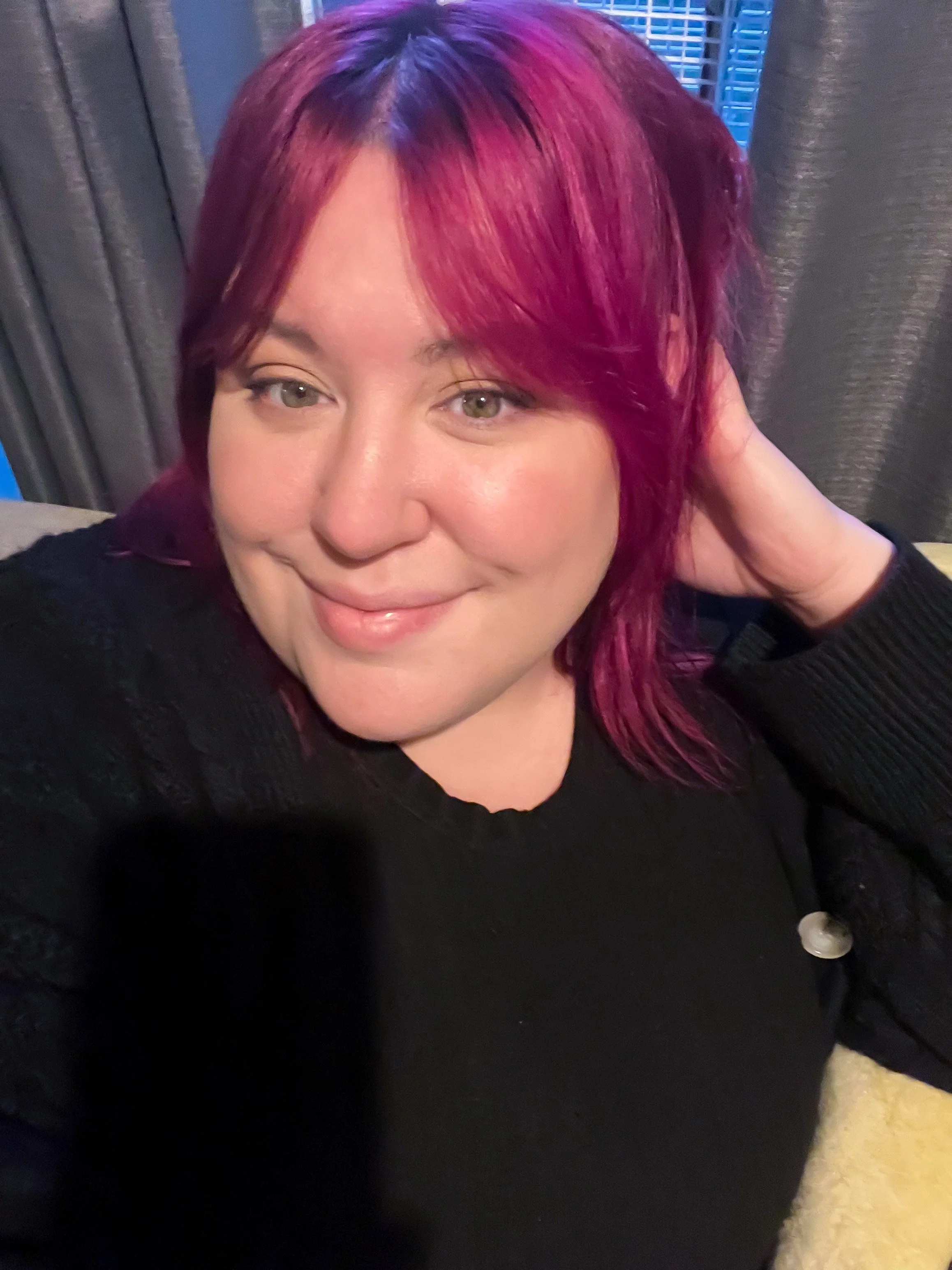 A woman with shoulder-length pink and purple hair smiling at the camera, wearing a black top with textured sleeves, indoors with dark curtains and a window behind her.