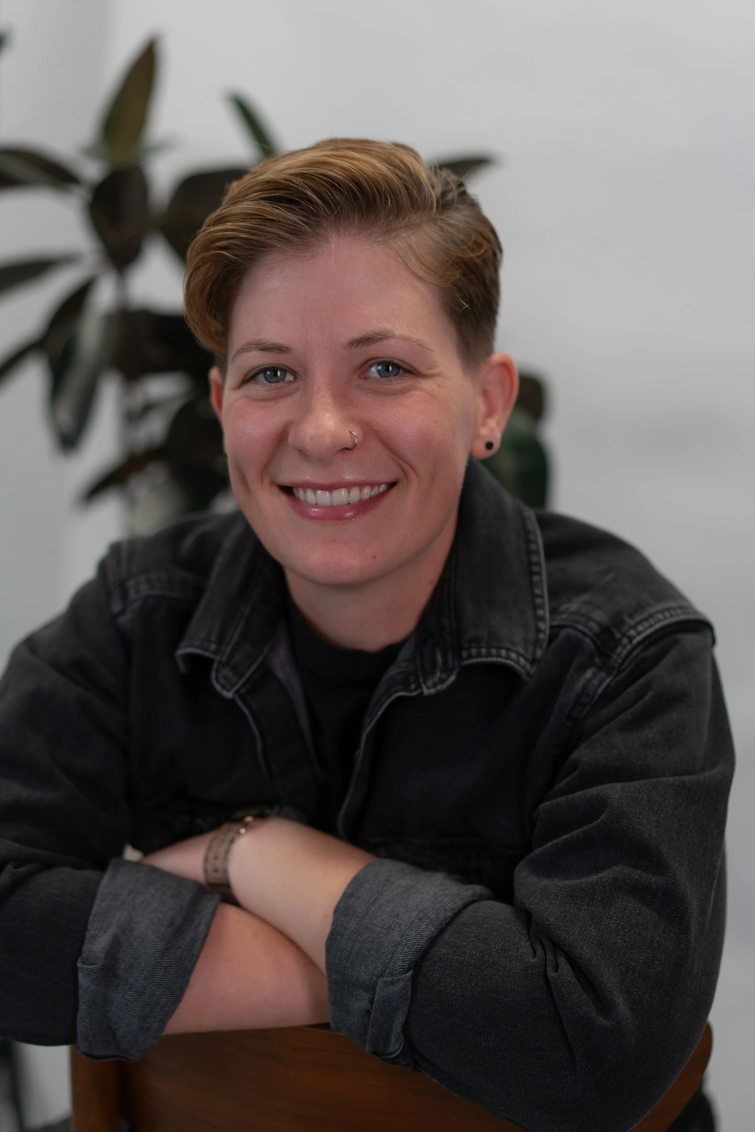 A smiling person with short, styled hair, wearing a black denim jacket, with arms crossed on a wooden chair, and a large plant in the background.
