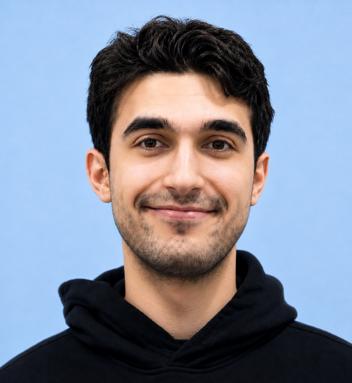 Close-up of a young man with dark hair, light skin, smiling, wearing a black hoodie, against a light blue background.