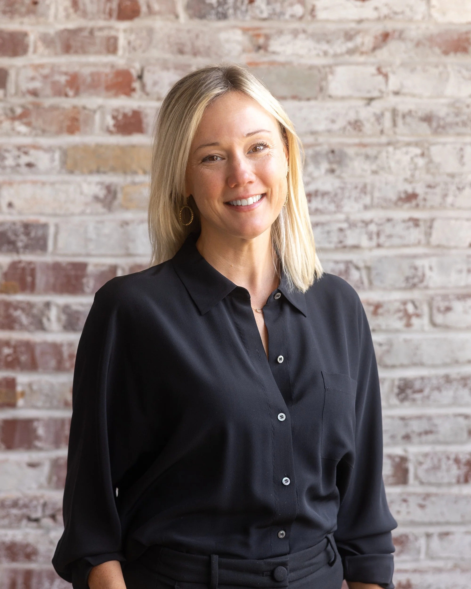 Portrait of a smiling woman with light-colored hair, wearing a dark shirt and a necklace, with a dark background.