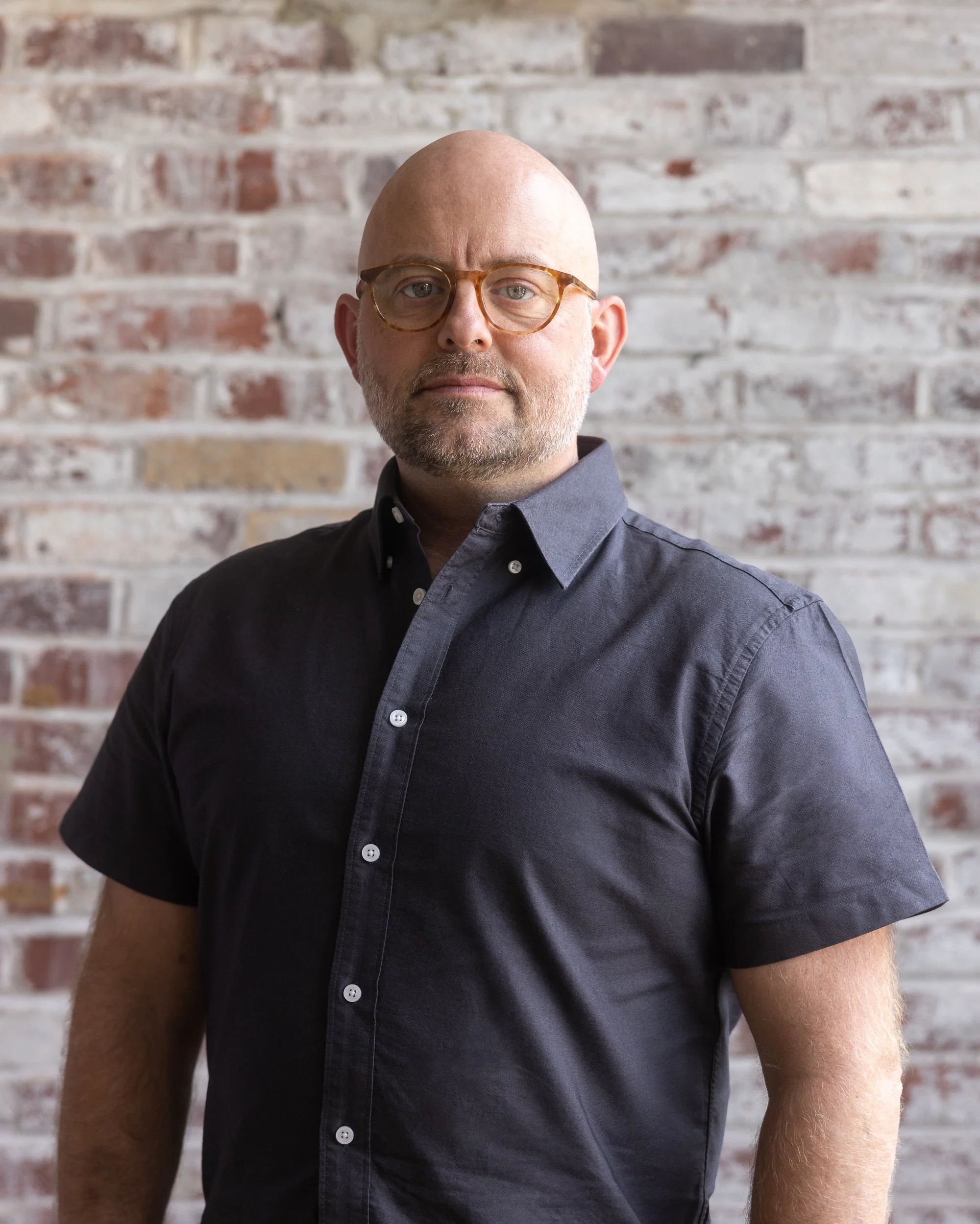 Close-up of a man with a bald head and a beard, wearing a black shirt, looking at the camera against a plain light background.