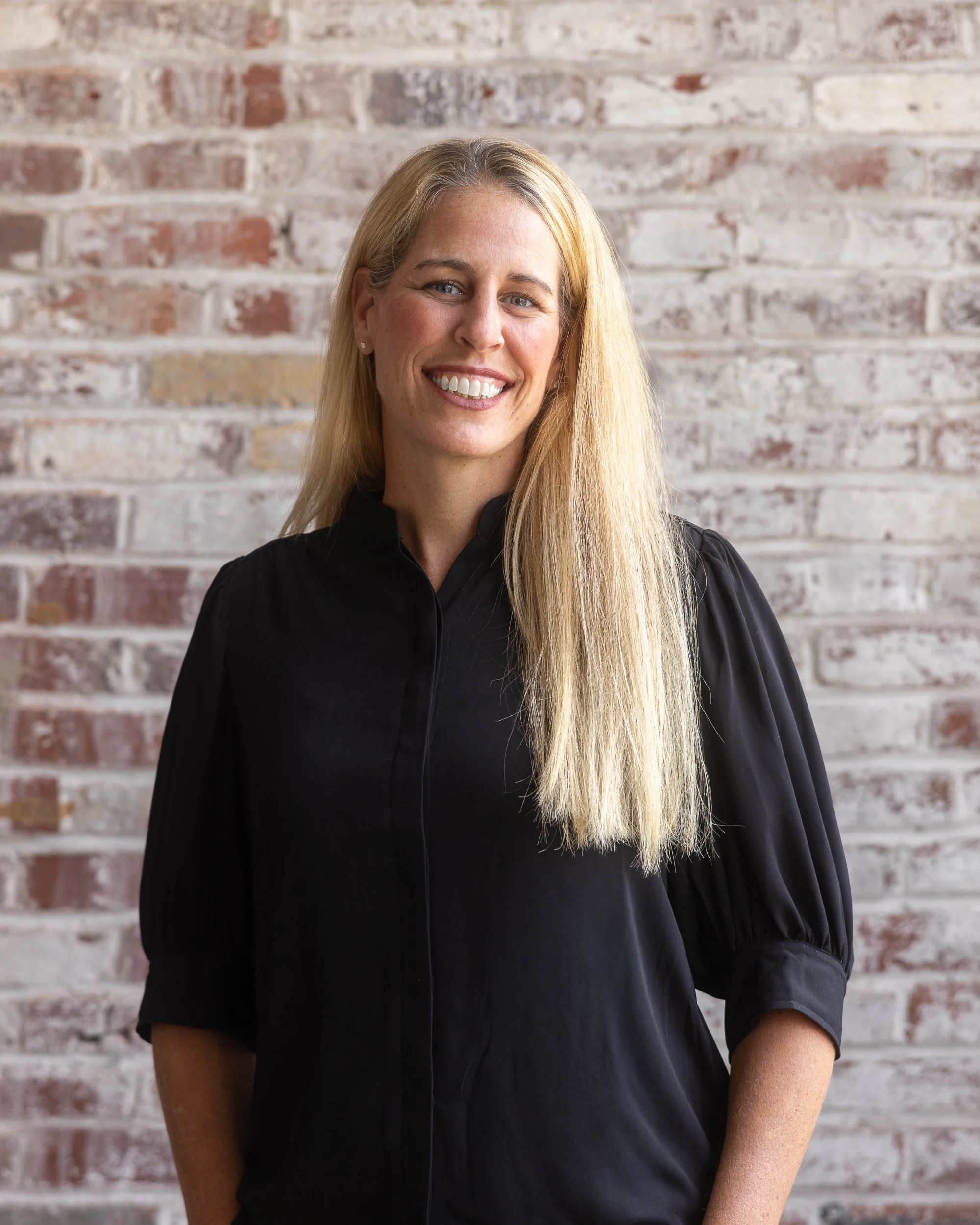 Photo of a woman smiling while looking out a window, with long blonde hair and wearing pearl earrings.