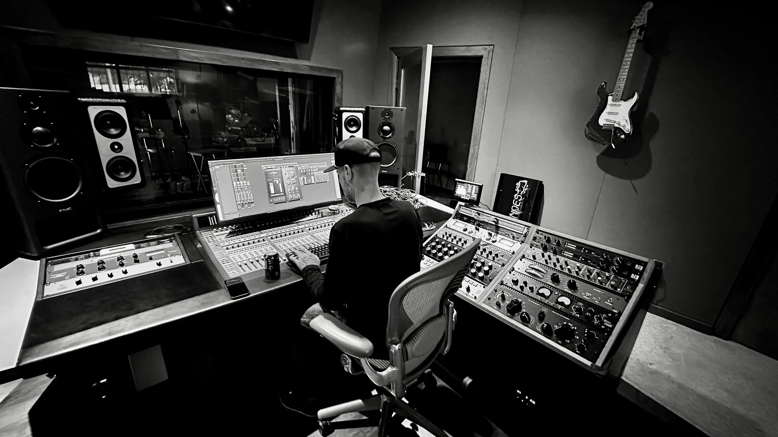 A person in a black cap and shirt working in a recording studio, surrounded by professional audio equipment, large speakers, a guitar hanging on the wall, and a mixing console.