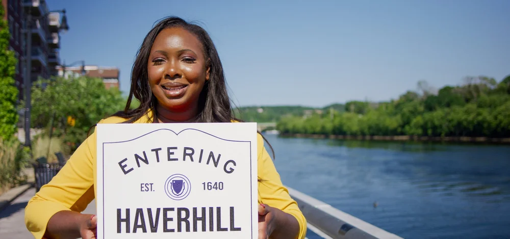 Eunice holding an Entering Haverhill sign beside the river