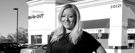 A smiling woman standing outside a building with the IN-N-OUT sign, in front of parked cars.