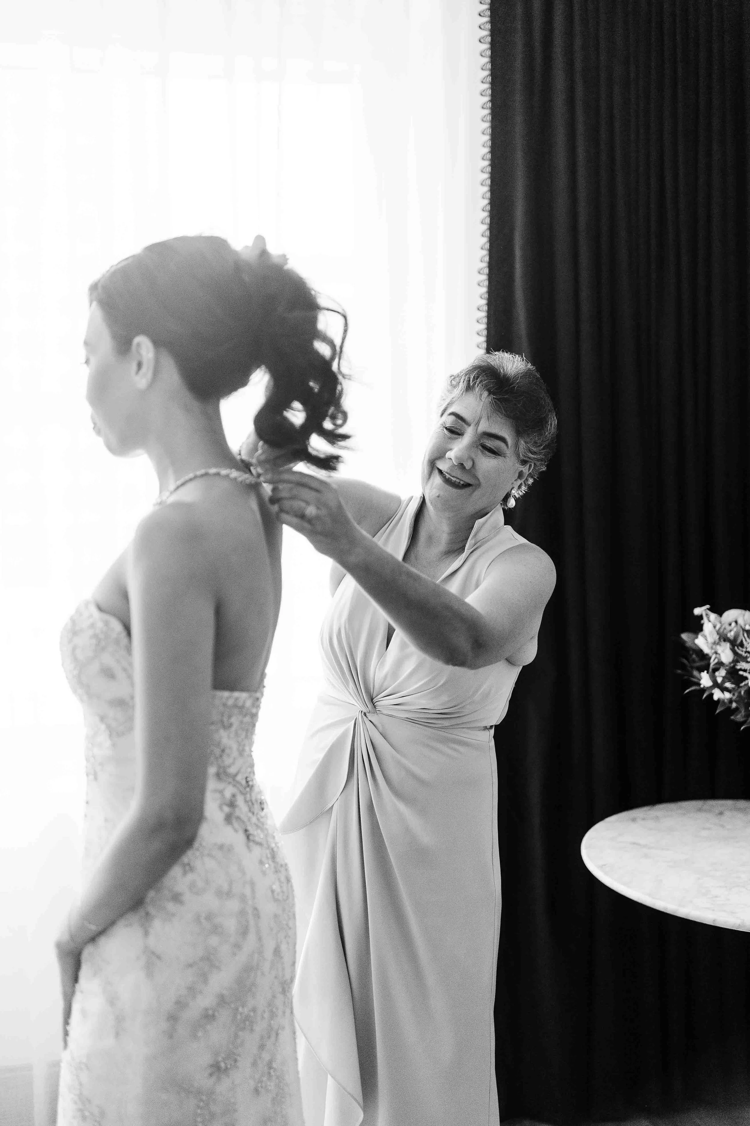 A woman helps another, dressed in a wedding gown, fasten a necklace in a softly lit room—captured beautifully by a Kansas City wedding photographer. Mother and daughter wedding photos Hotel Kansas City