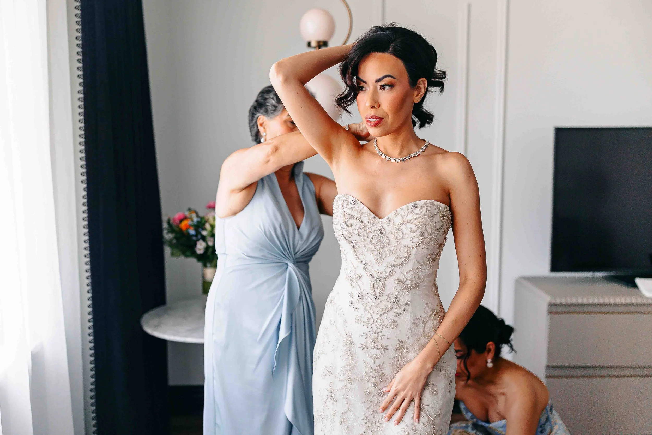 A bride in an ornate strapless gown adjusts her hair while two women help her get ready in a bright room—perfect for KC wedding photography.
