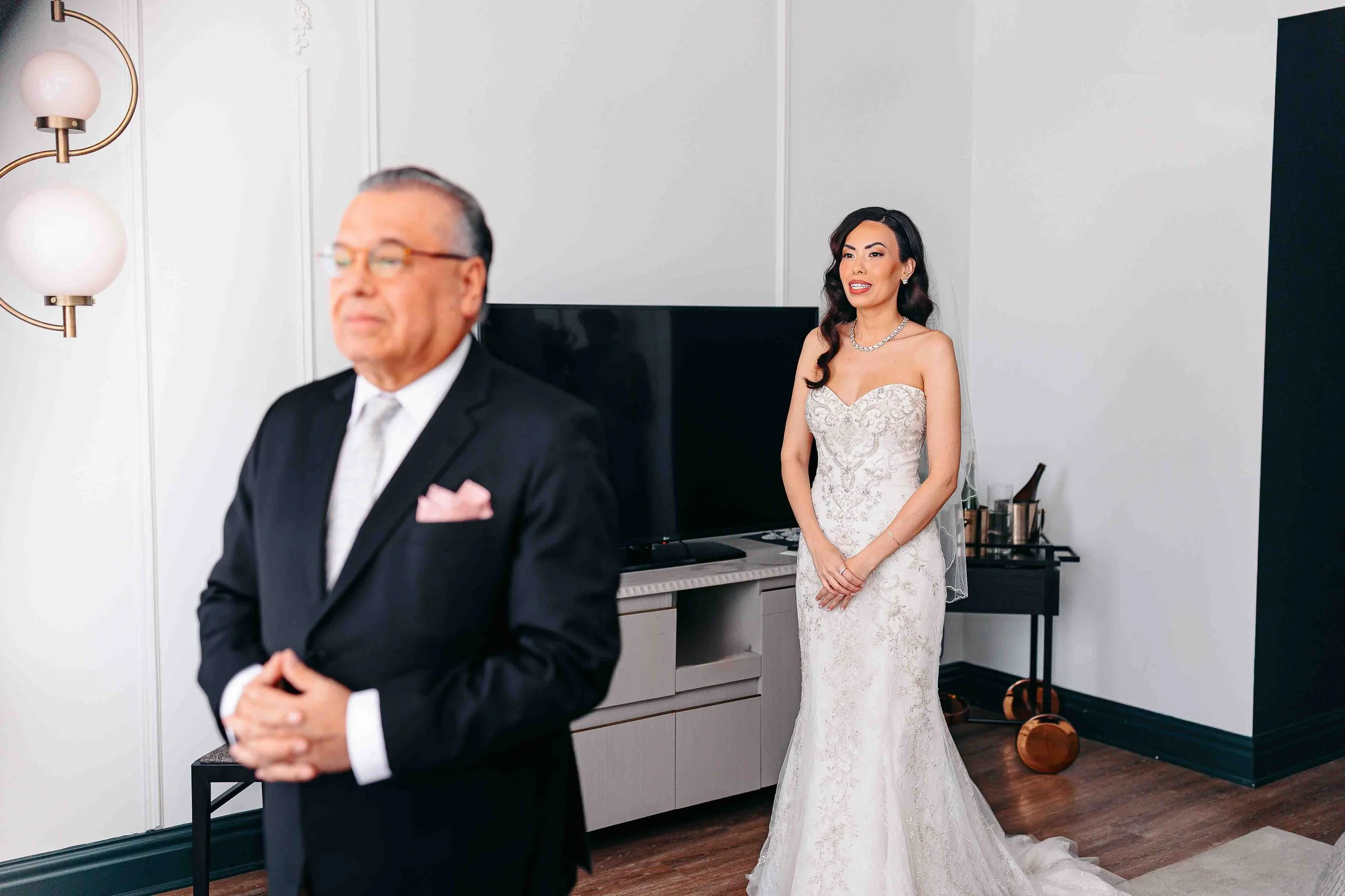 A bride in a white strapless gown stands smiling behind an older man in a suit, inside a modern room—captured by Humbly Hewn Photography.