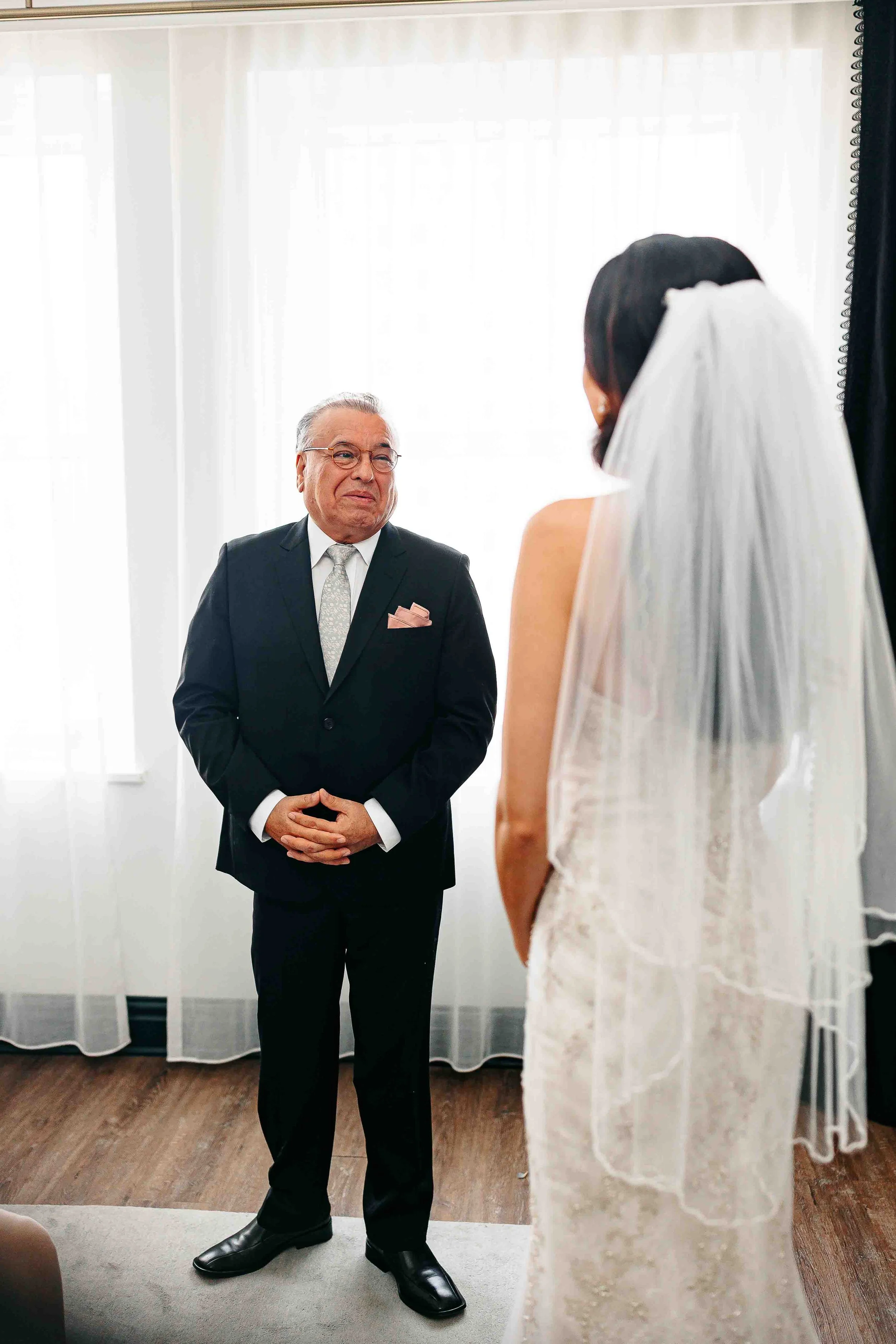 An older man in a suit smiles warmly at a bride in a white dress and veil, captured in bright light by KC wedding photography.