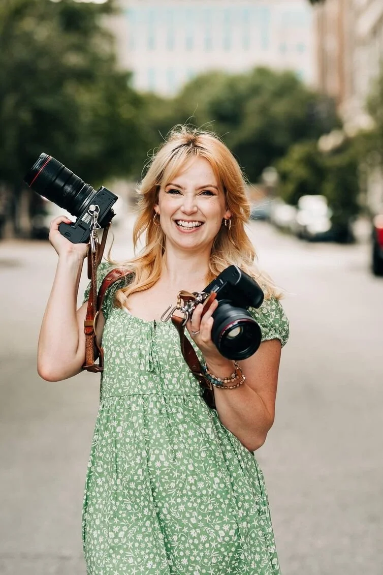 A woman in a green floral dress smiles and holds two cameras, embodying the charm of a Kansas City wedding photographer amidst trees and blurred buildings.