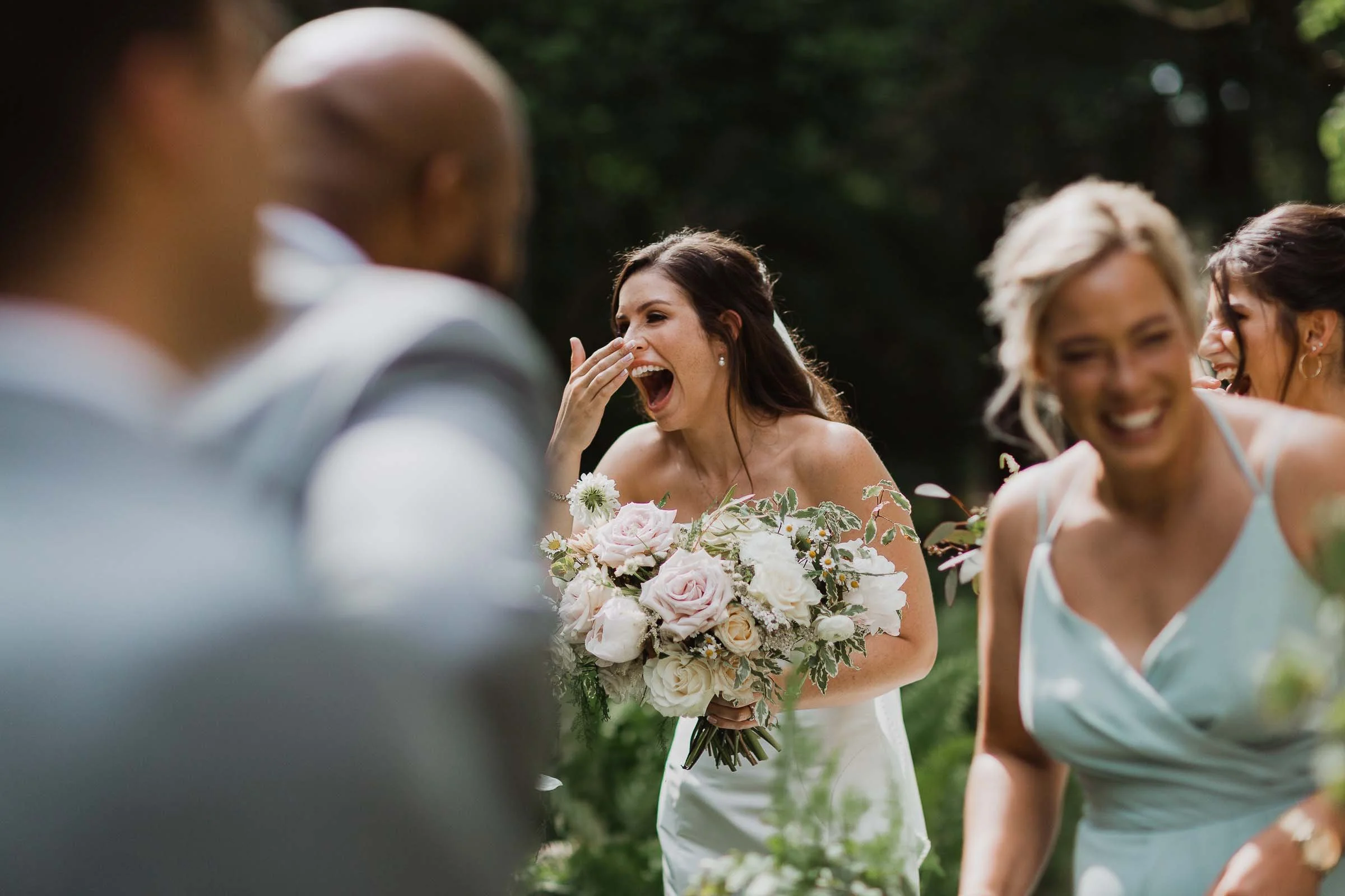A bride laughs with bridesmaids and groomsmen outdoors in light attire at Westcroft Gardens, captured by a talented Grosse Ile wedding photographer on a sunny day.