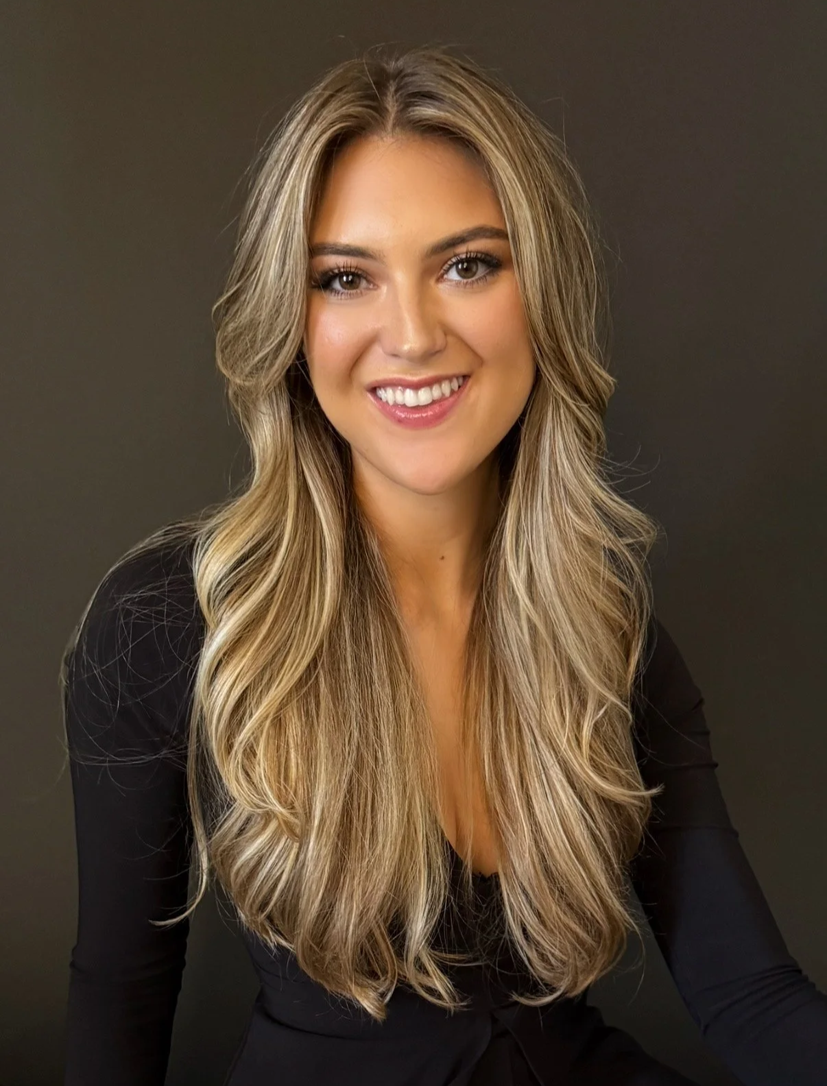 Portrait of a smiling woman with long, wavy blonde hair, wearing a black top against a dark background.