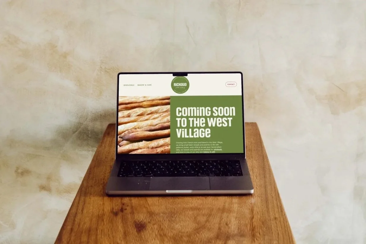 Laptop on wooden table displaying a webpage for a bakery called Richaud Petite Bakery, with a green and white design, and a message 'COMING SOON TO THE WEST VILLAGE'.