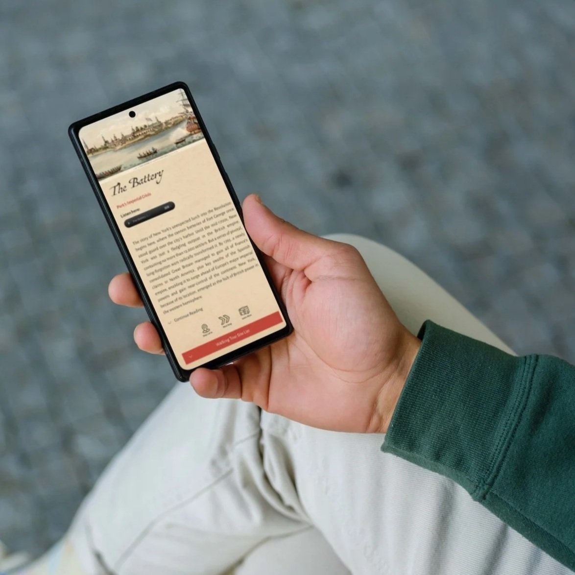 Person holding a smartphone with a webpage titled 'The Battery', featuring a historical image at the top, sitting outdoors on a gray surface.