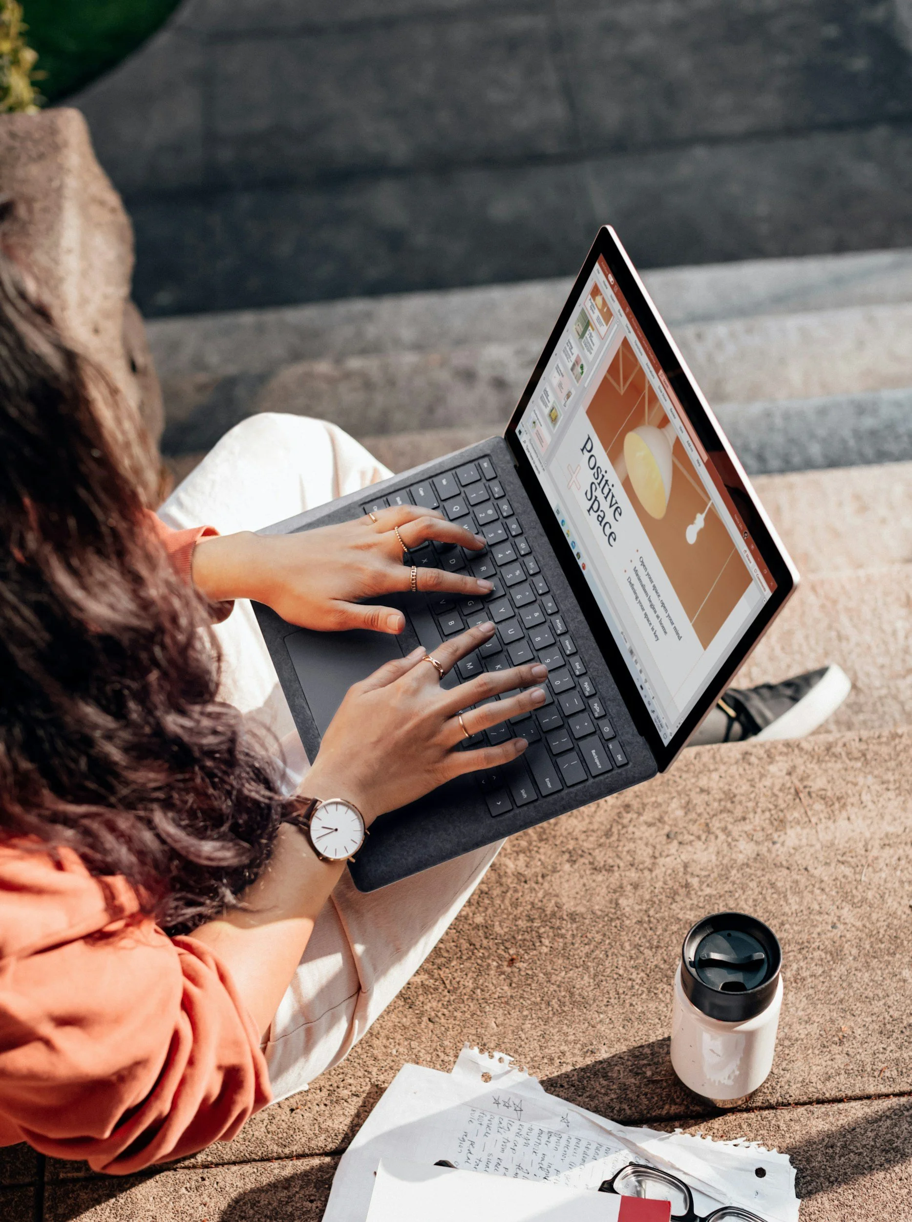 Person sitting outdoors using a laptop with a presentation on screen. Paperwork, a water bottle, and a coffee cup are nearby.