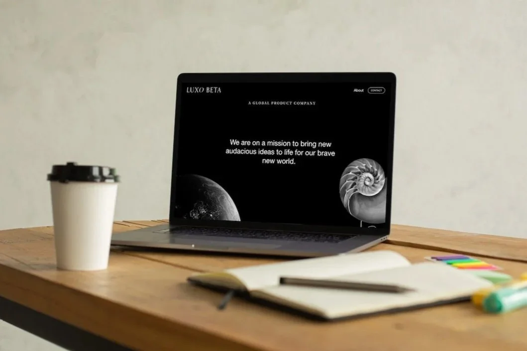 A laptop on a wooden desk displaying a dark-themed website for Luxo Beta with images of a moon and a shell. To the left of the laptop is a disposable coffee cup with a black lid. In front of the laptop, there is an open notebook and a pen.