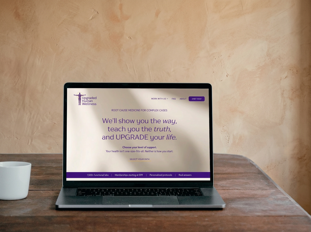 Laptop on a wooden table displaying the Upgraded Human Wellness website with purple text and a logo of a person with outstretched arms. Next to the laptop is a white mug, and the background features a beige wall.