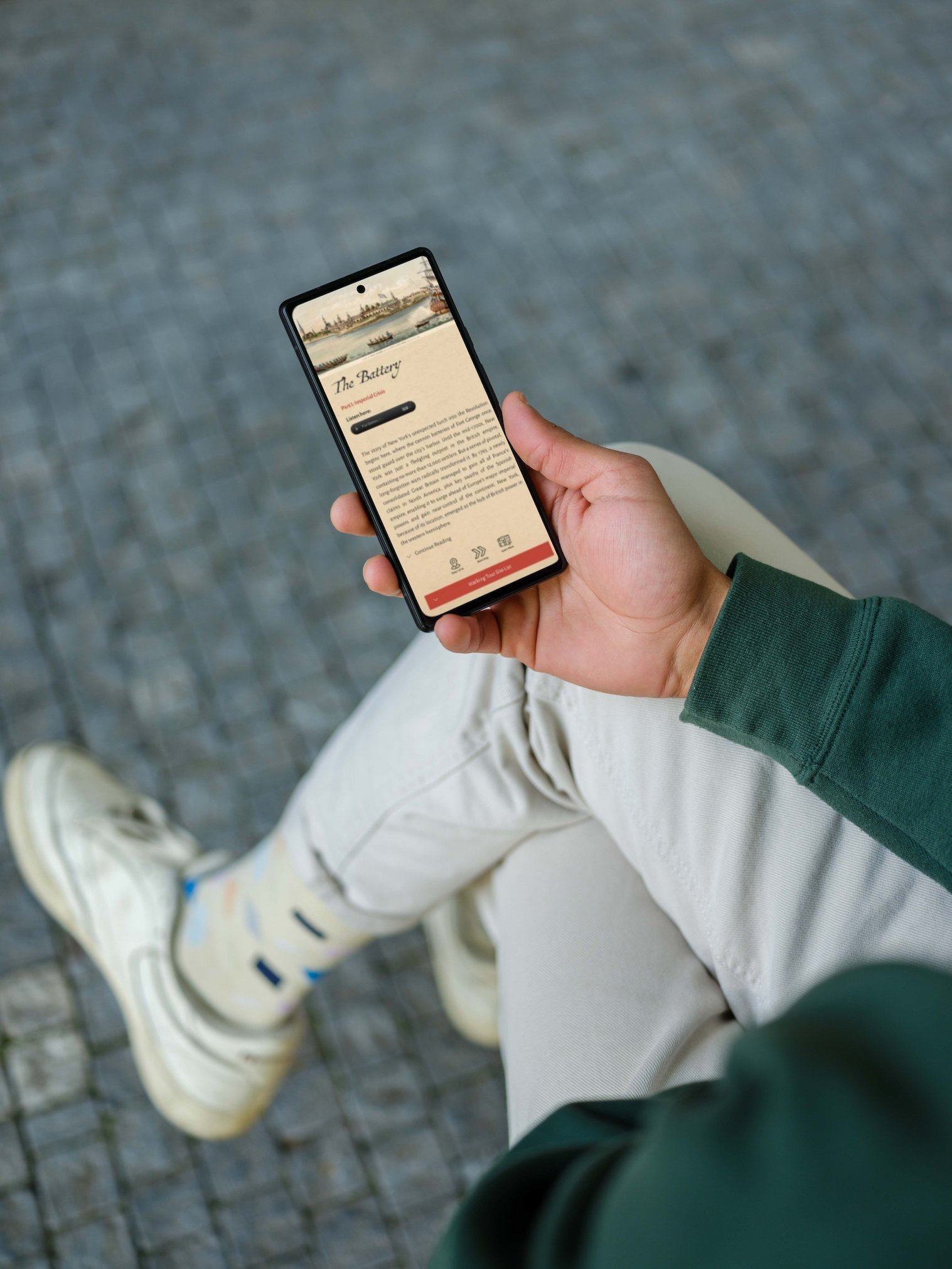 Person holding a smartphone viewing the NYC Revolutionary Trail walking tour visible on the screen, sitting outdoors on a cobblestone surface, wearing light-colored pants, white sneakers, and a dark green jacket.