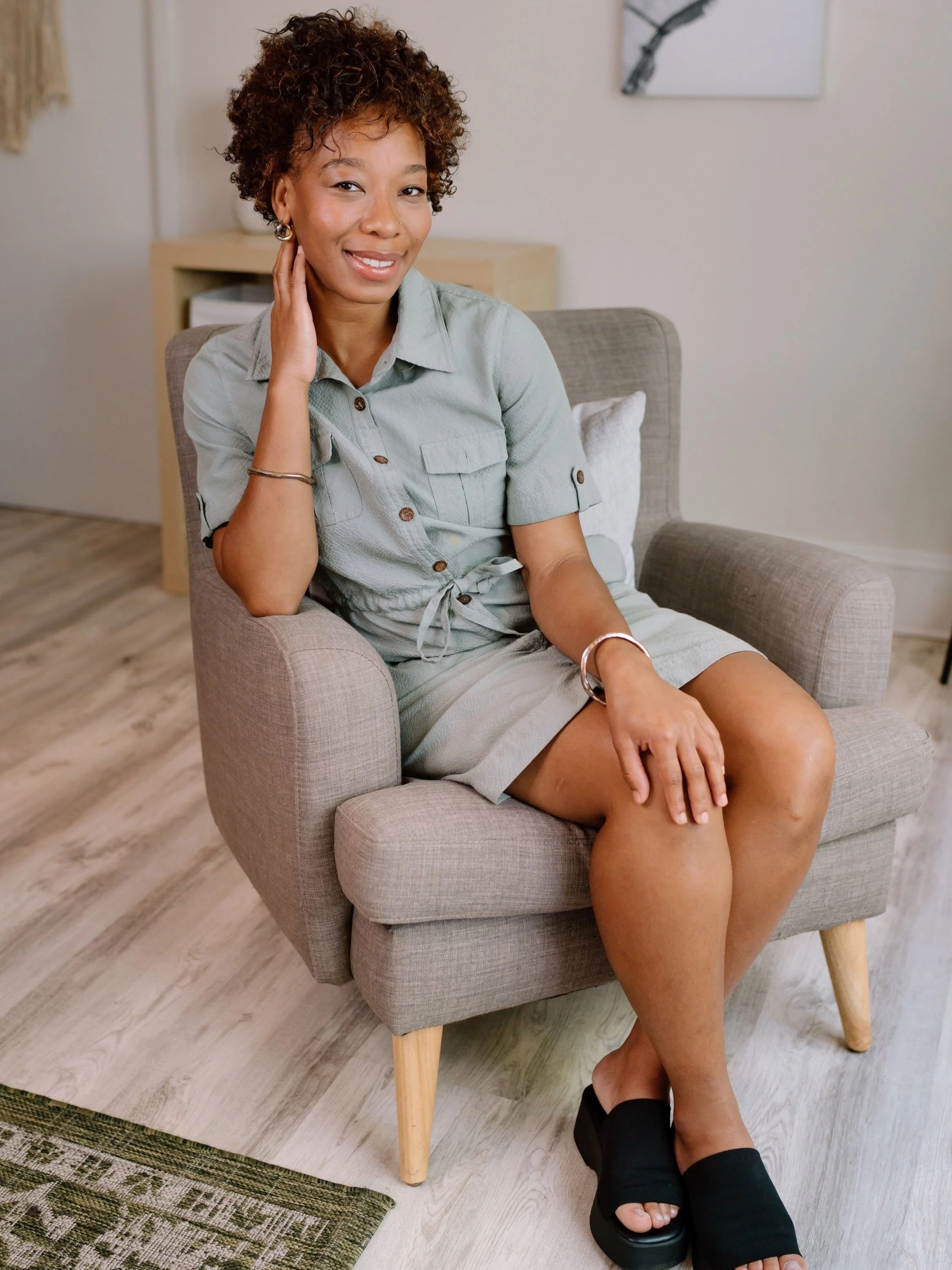 Woman sitting in a gray armchair in a casual setting, wearing a light green dress and black sandals. The room has wooden flooring and neutral decor.