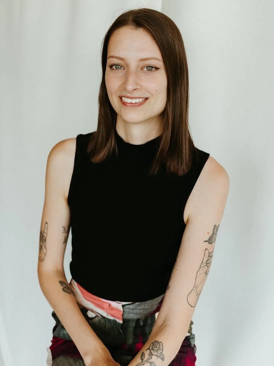 A person with shoulder-length brown hair, wearing a sleeveless black top. They have tattoos on their arms and are smiling. The background is white.