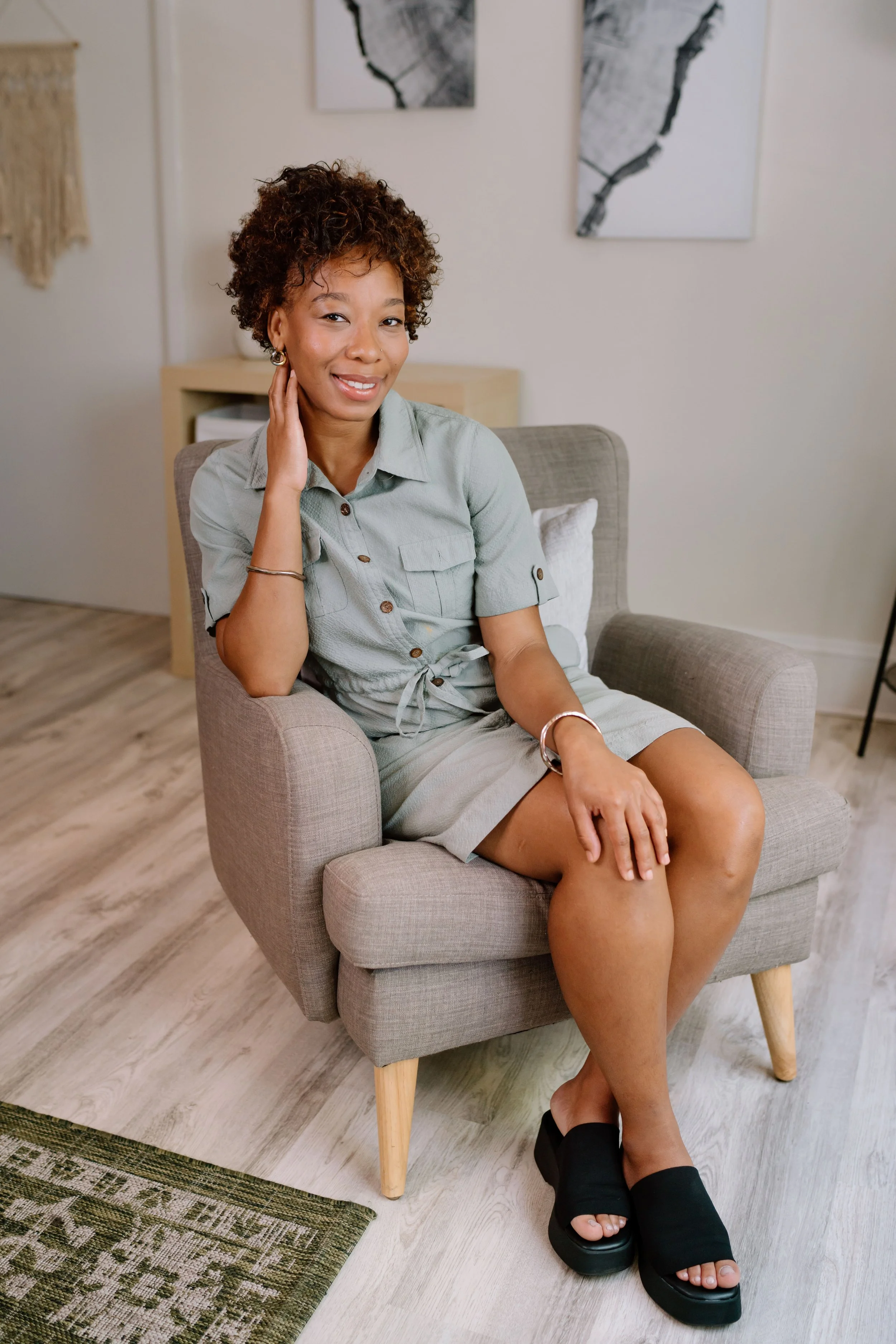 Woman sitting on a gray chair wearing a light green dress and black open-toe heels, smiling with a hand on her neck.