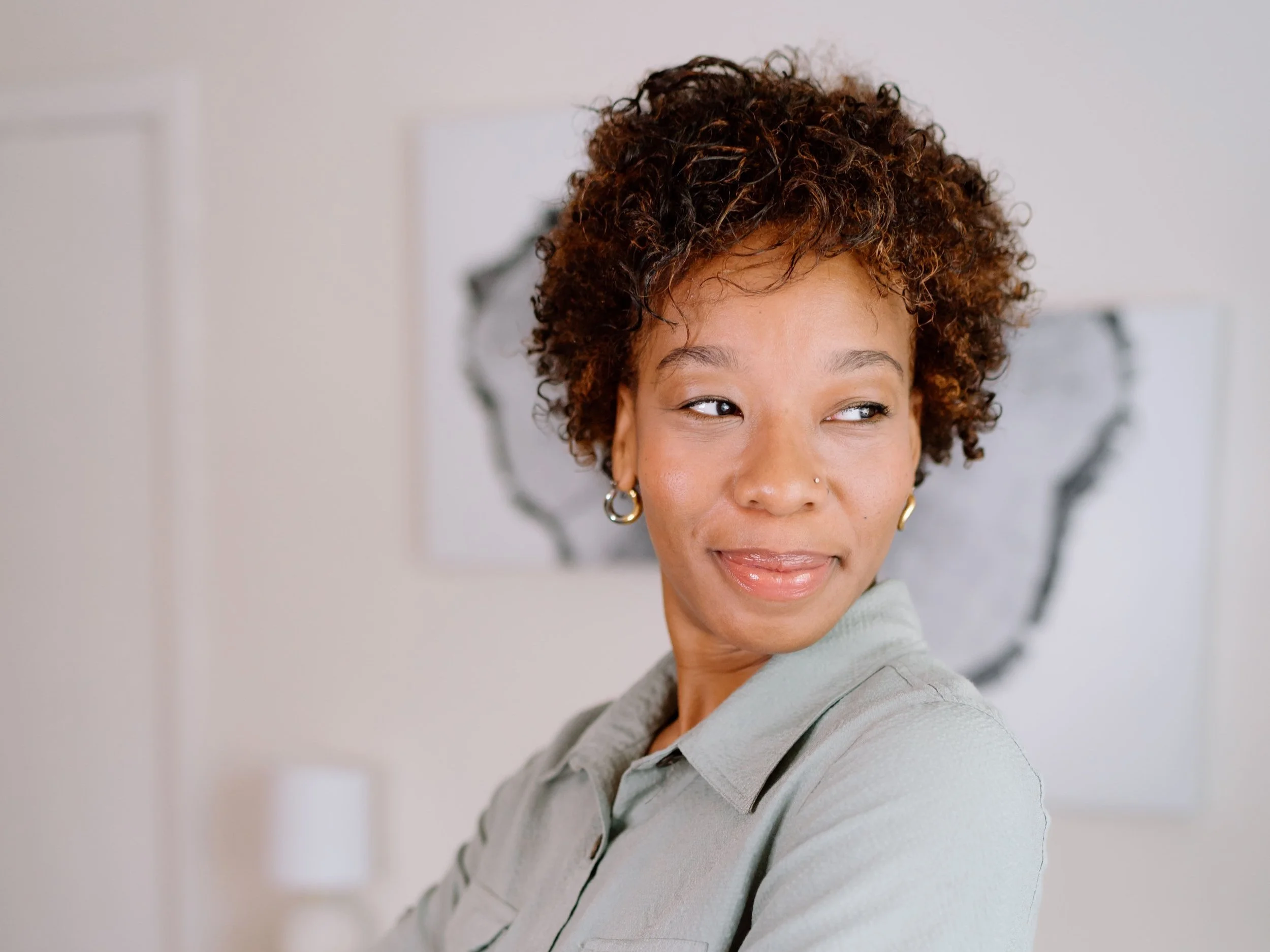 Person with short curly hair looking sideways, wearing a light shirt and hoop earrings, with abstract art in the background.