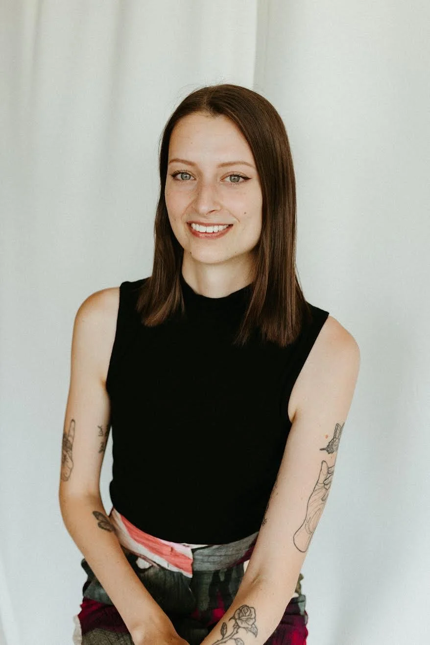 Person smiling with tattoos on arms, wearing a black top and patterned skirt, against a light background.