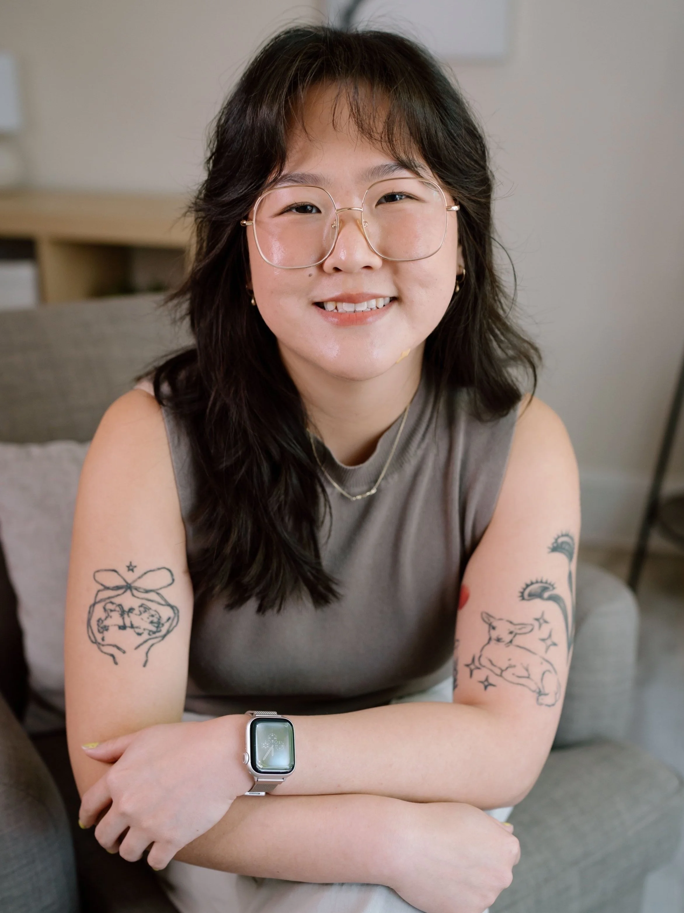 Person with glasses and tattoos on both arms, wearing a smartwatch, seated on a couch, smiling.