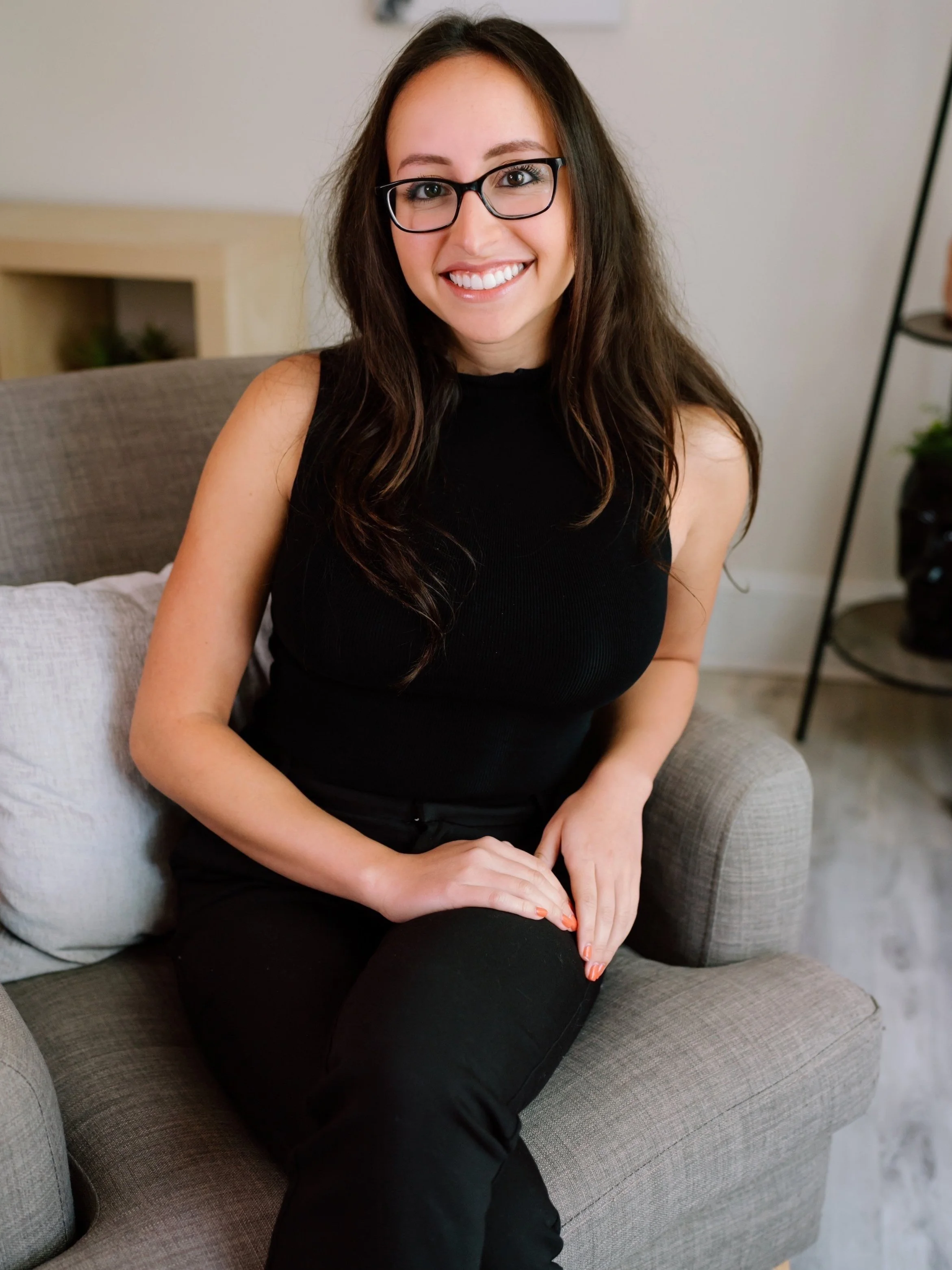Smiling person with glasses sitting on a gray armchair indoors, wearing a black sleeveless top and pants.