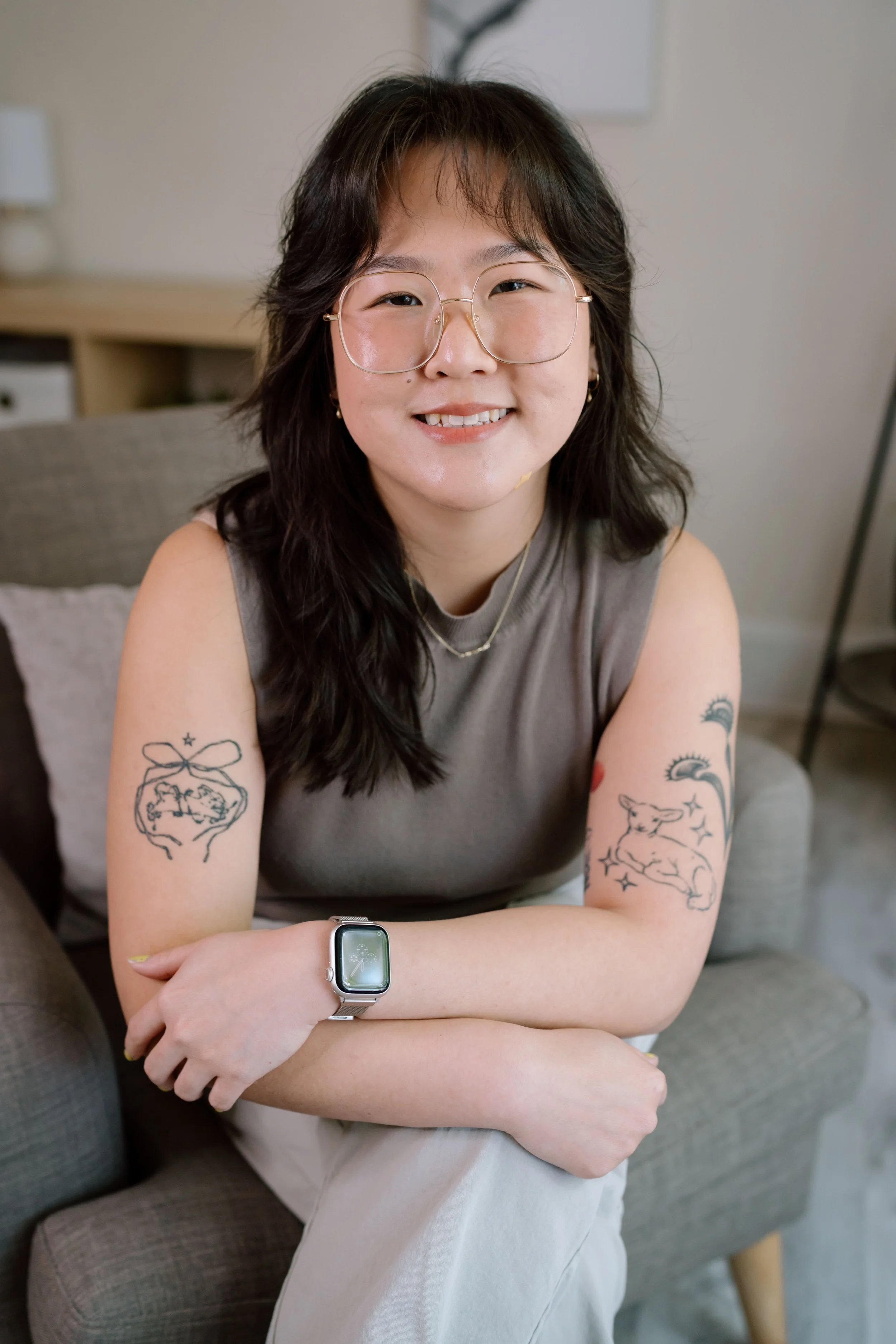 Person with glasses, tattoos on both arms, wearing a sleeveless top, sitting on a gray chair, and wearing a smartwatch.