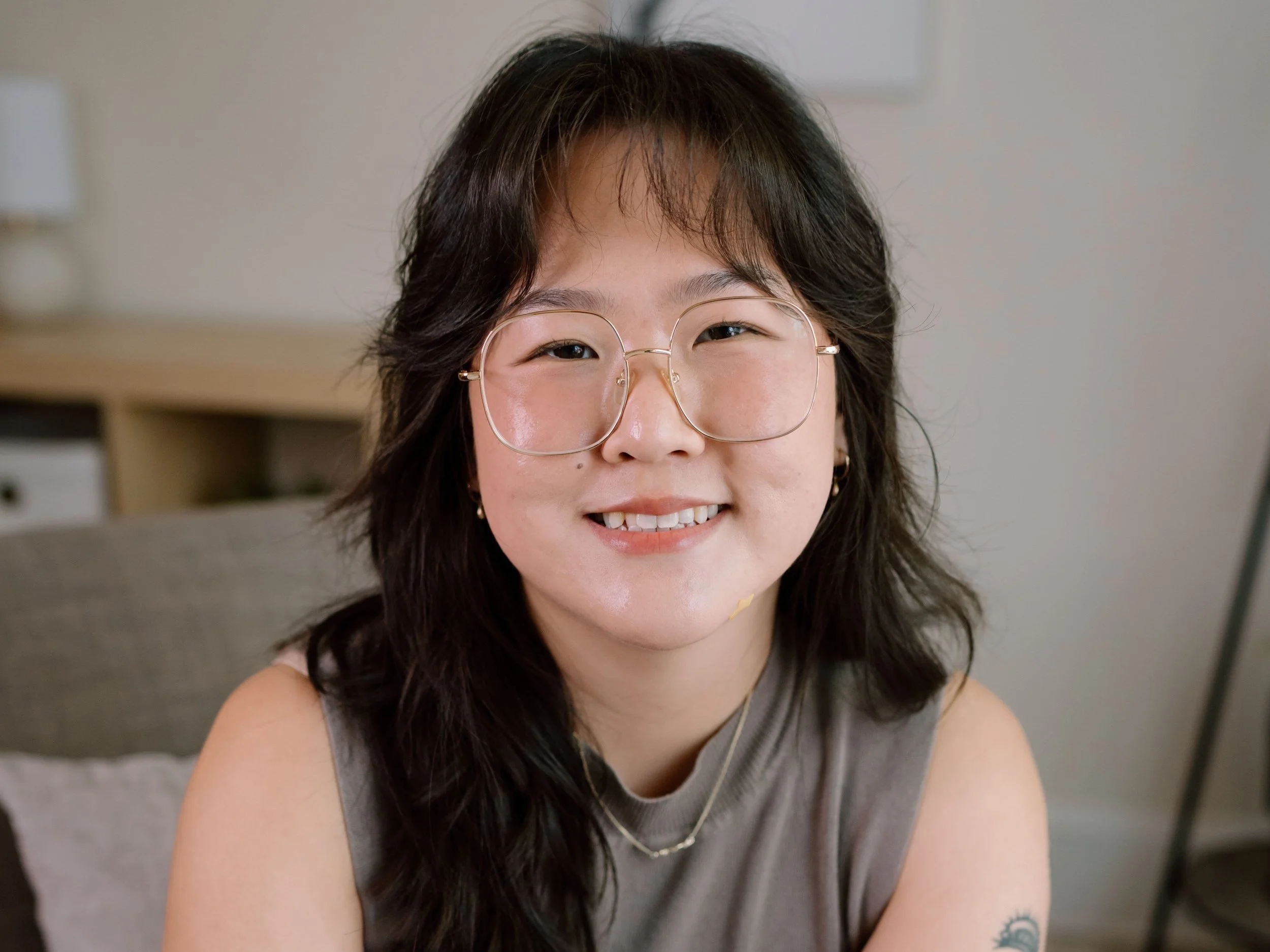 Person with glasses smiling, wearing a gray sleeveless shirt, indoors.