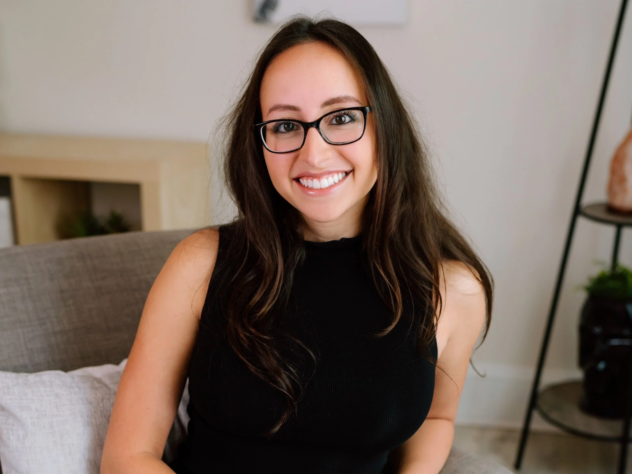 Woman with long brown hair and glasses smiling, sitting on a gray sofa.