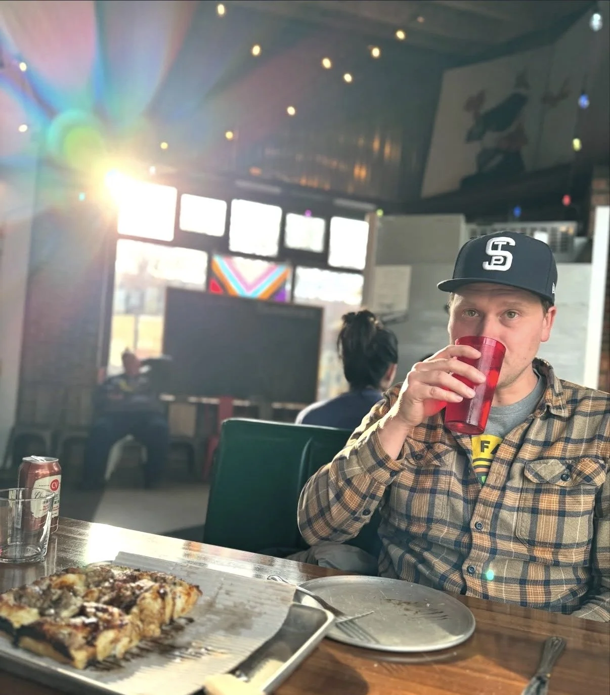 Man wearing a black baseball cap with white logo, plaid shirt, and gray t-shirt, drinking from a red cup at a restaurant with pizza on the table, sunlight coming through windows and other patrons in the background.
