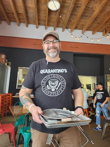 Man with a beard and glasses, wearing a black Quarantino's t-shirt, holding a tray of food, standing inside a restaurant with wooden ceiling, colorful chairs, and other people in the background.