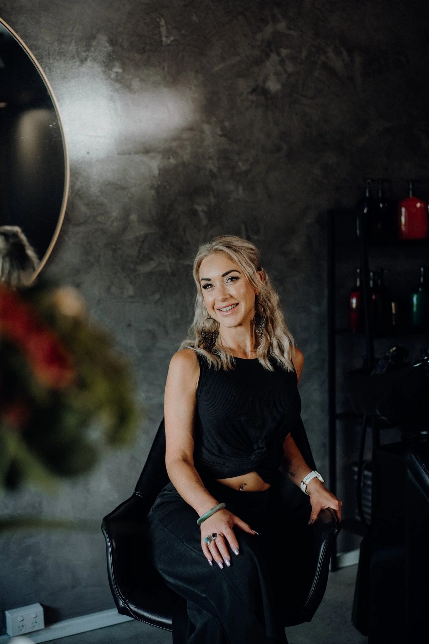 A smiling woman with wavy blonde hair sitting on a black chair in a dark, modern interior with a gray textured wall. She is wearing a black sleeveless top, black pants, and accessories including rings, earrings, a bracelet, and a watch. There are shelves with red and green bottles in the background.