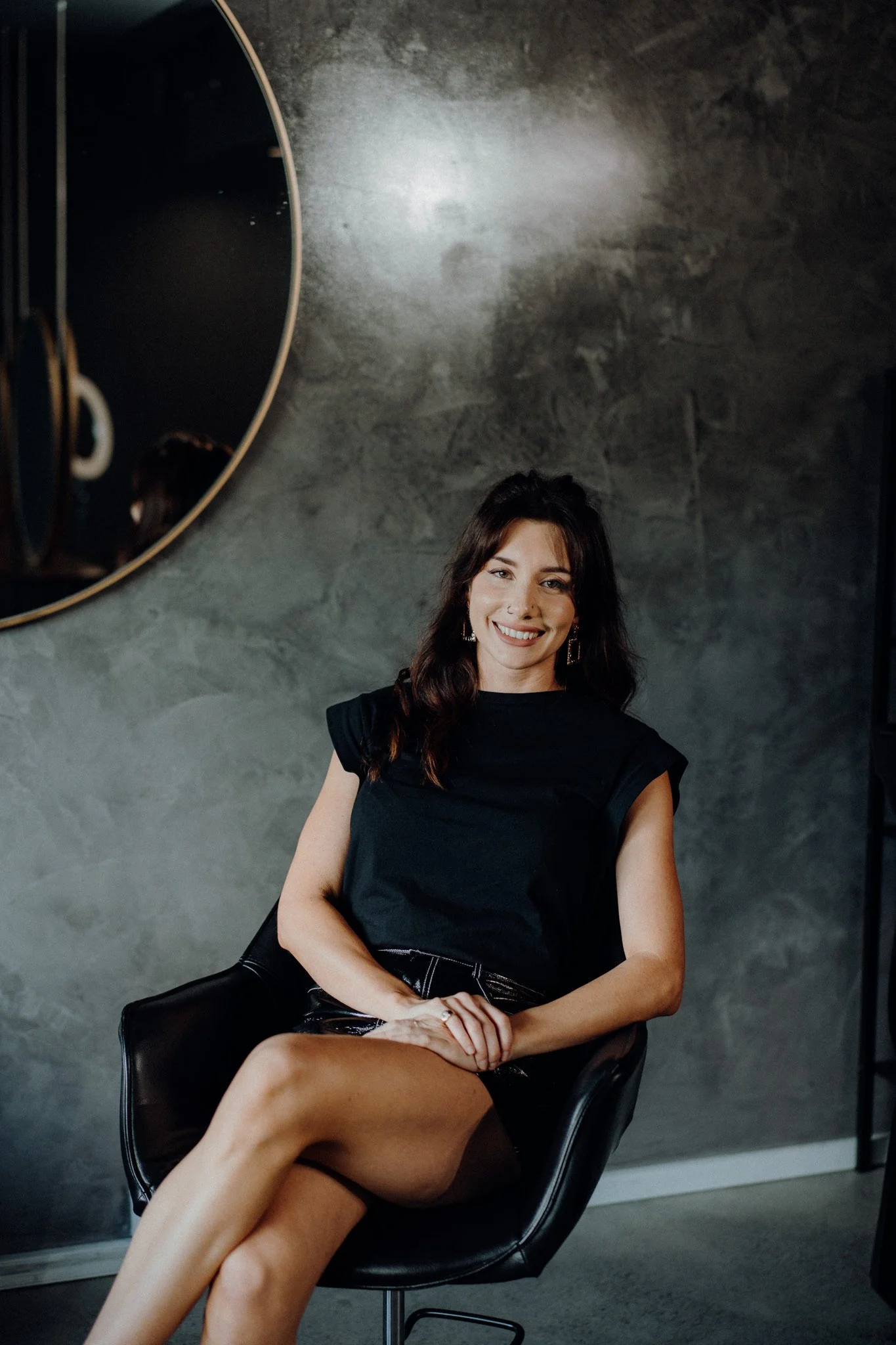 A woman with shoulder-length dark hair and a big smile sitting on a black chair against a textured gray wall with a round mirror reflecting part of her face and a darker room.