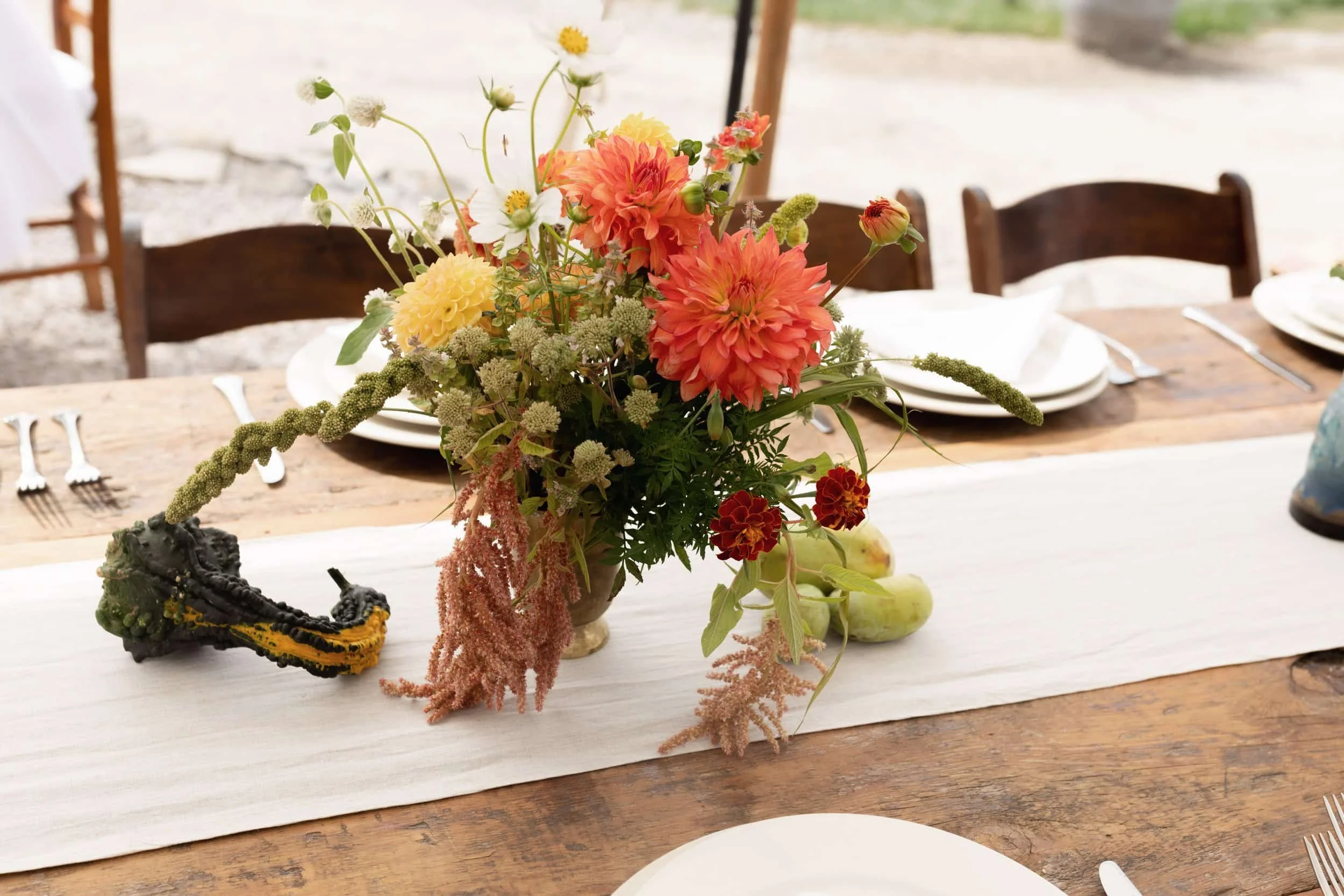 A wooden dining table with a white table runner, set with white plates and silverware, featuring a floral centerpiece with orange, yellow, white, and green flowers in a brown vase. There is a decorative black and yellow gourd and two green gourds on the table.