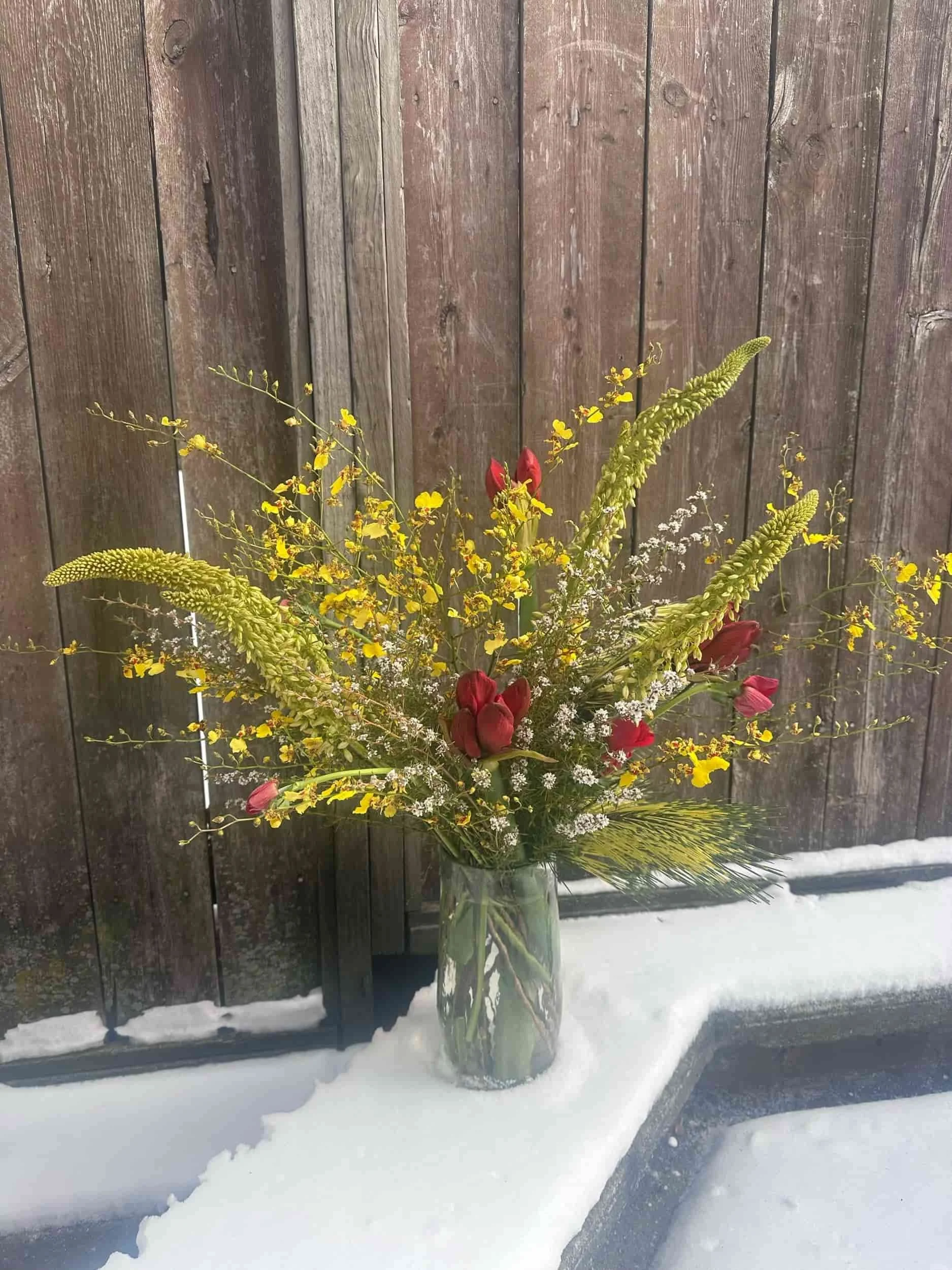 A colorful flower arrangement in a glass vase placed on snow in front of a wooden fence.