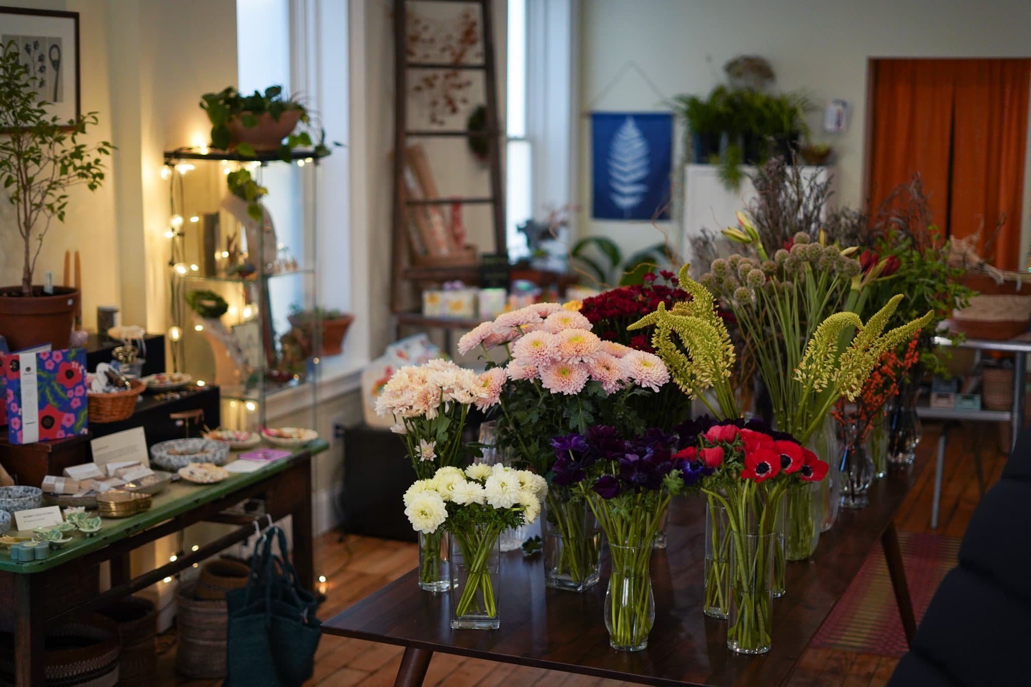 A table filled with various colorful flowers in glass vases inside a cozy indoor space, with shelves, plants, and decorative items in the background.